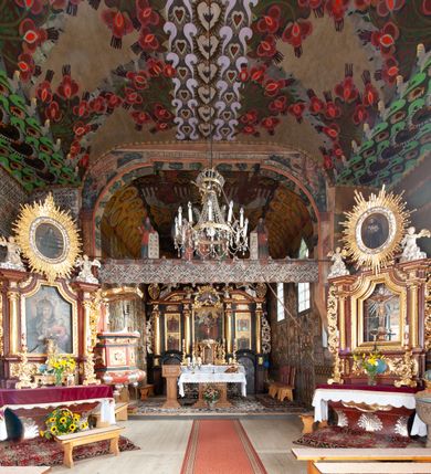 Zdjęcie nr 8: Kościół drewniany, orientowany, składający się z babińca, nawy głównej z przylegającą do niej od północy kaplicą boczną i kruchtą od południa oraz prezbiterium z zakrystią od północy. Prezbiterium i nawa w konstrukcji zrębowej, wieża, soboty i kruchta w słupowej. Kościół jednonawowy, nawa prawie kwadratowa, prezbiterium prostokątne, zamknięte prosto, oddzielone ścianą tęczową. Otwór tęczowy zamknięty łukiem dwuramiennym z biegnącą w poprzek grubą belką. Ściany gładkie, pokryte dekoracją malarską imitującą poziomo ułożone belki z uszczelniającymi je kiczkami, scenami figuralnymi, zacheuszkami i pasami ornamentalnymi. W nawie od zachodu chór muzyczny wsparty na dwóch, profilowanych słupach, z parapetem wybrzuszonym pośrodku; z dekoracją malarską o motywach geometryczno-roślinnych. Nawa i prezbiterium naryte pozornym sklepieniem z pułapami lekko zaokrąglonymi po bokach. Na całości przekrycia dekoracja malarska. W prezbiterium są to postacie aniołów wśród stylizowanych kwiatów na stropie adorujących monogram maryjny, nawę dekorują motywy roślinne. Anioły zdobią również sklepienie kaplicy. Strop babińca oraz fragmenty prezbiterium pokrywa oryginalna dekoracja patronowa złożona z motywów geometrycznych oraz figuralnych. W nawie głównej trzy okna, w prezbiterium i babińcu po dwa – od południa, kolejne do zakrystii; zamknięte łukiem dwuspadowym. Główne wejście od zachodu, drugie przez kruchtę od południa, trzecie do zakrystii. Portal południowy ostrołukowy, dekorowany ornamentem sznurowym, portal do zakrystii zamknięty łukiem w ośli grzbiet. Od zachodu kwadratowa wieża o pochyłych ścianach, z nadwieszoną izbicą, zwieńczona czteropołaciowym, ostrosłupowym hełmem gontowym. Izbica wieży oszalowana dookoła deskami zakończonymi u dołu ozdobnie wyrzynaną koronką. Od południa i wschodu soboty, czyli rodzaj podcieni,  w pozostałych miejscach gontowy kołnierz. Ściany świątyni oraz kaplicy są od zewnątrz szalowane deskami, wieża oraz przedsionek obite są gontem. Dachy i zadaszenia sobot pokryte gontem, kopuła kaplicy oraz wieżyczka na sygnaturkę pokryta blachą. 