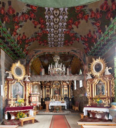 Zdjęcie nr 6: Kościół drewniany, orientowany, składający się z babińca, nawy głównej z przylegającą do niej od północy kaplicą boczną i kruchtą od południa oraz prezbiterium z zakrystią od północy. Prezbiterium i nawa w konstrukcji zrębowej, wieża, soboty i kruchta w słupowej. Kościół jednonawowy, nawa prawie kwadratowa, prezbiterium prostokątne, zamknięte prosto, oddzielone ścianą tęczową. Otwór tęczowy zamknięty łukiem dwuramiennym z biegnącą w poprzek grubą belką. Ściany gładkie, pokryte dekoracją malarską imitującą poziomo ułożone belki z uszczelniającymi je kiczkami, scenami figuralnymi, zacheuszkami i pasami ornamentalnymi. W nawie od zachodu chór muzyczny wsparty na dwóch, profilowanych słupach, z parapetem wybrzuszonym pośrodku; z dekoracją malarską o motywach geometryczno-roślinnych. Nawa i prezbiterium naryte pozornym sklepieniem z pułapami lekko zaokrąglonymi po bokach. Na całości przekrycia dekoracja malarska. W prezbiterium są to postacie aniołów wśród stylizowanych kwiatów na stropie adorujących monogram maryjny, nawę dekorują motywy roślinne. Anioły zdobią również sklepienie kaplicy. Strop babińca oraz fragmenty prezbiterium pokrywa oryginalna dekoracja patronowa złożona z motywów geometrycznych oraz figuralnych. W nawie głównej trzy okna, w prezbiterium i babińcu po dwa – od południa, kolejne do zakrystii; zamknięte łukiem dwuspadowym. Główne wejście od zachodu, drugie przez kruchtę od południa, trzecie do zakrystii. Portal południowy ostrołukowy, dekorowany ornamentem sznurowym, portal do zakrystii zamknięty łukiem w ośli grzbiet. Od zachodu kwadratowa wieża o pochyłych ścianach, z nadwieszoną izbicą, zwieńczona czteropołaciowym, ostrosłupowym hełmem gontowym. Izbica wieży oszalowana dookoła deskami zakończonymi u dołu ozdobnie wyrzynaną koronką. Od południa i wschodu soboty, czyli rodzaj podcieni,  w pozostałych miejscach gontowy kołnierz. Ściany świątyni oraz kaplicy są od zewnątrz szalowane deskami, wieża oraz przedsionek obite są gontem. Dachy i zadaszenia sobot pokryte gontem, kopuła kaplicy oraz wieżyczka na sygnaturkę pokryta blachą. 