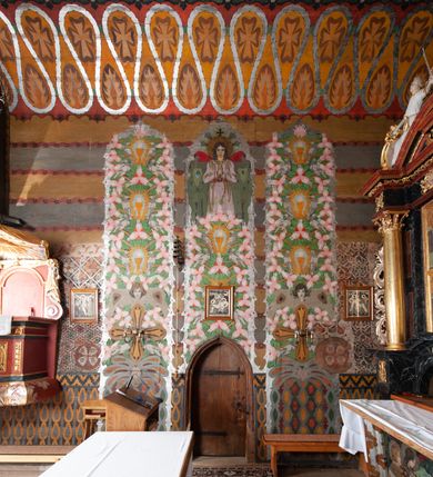 Zdjęcie nr 14: Kościół drewniany, orientowany, składający się z babińca, nawy głównej z przylegającą do niej od północy kaplicą boczną i kruchtą od południa oraz prezbiterium z zakrystią od północy. Prezbiterium i nawa w konstrukcji zrębowej, wieża, soboty i kruchta w słupowej. Kościół jednonawowy, nawa prawie kwadratowa, prezbiterium prostokątne, zamknięte prosto, oddzielone ścianą tęczową. Otwór tęczowy zamknięty łukiem dwuramiennym z biegnącą w poprzek grubą belką. Ściany gładkie, pokryte dekoracją malarską imitującą poziomo ułożone belki z uszczelniającymi je kiczkami, scenami figuralnymi, zacheuszkami i pasami ornamentalnymi. W nawie od zachodu chór muzyczny wsparty na dwóch, profilowanych słupach, z parapetem wybrzuszonym pośrodku; z dekoracją malarską o motywach geometryczno-roślinnych. Nawa i prezbiterium naryte pozornym sklepieniem z pułapami lekko zaokrąglonymi po bokach. Na całości przekrycia dekoracja malarska. W prezbiterium są to postacie aniołów wśród stylizowanych kwiatów na stropie adorujących monogram maryjny, nawę dekorują motywy roślinne. Anioły zdobią również sklepienie kaplicy. Strop babińca oraz fragmenty prezbiterium pokrywa oryginalna dekoracja patronowa złożona z motywów geometrycznych oraz figuralnych. W nawie głównej trzy okna, w prezbiterium i babińcu po dwa – od południa, kolejne do zakrystii; zamknięte łukiem dwuspadowym. Główne wejście od zachodu, drugie przez kruchtę od południa, trzecie do zakrystii. Portal południowy ostrołukowy, dekorowany ornamentem sznurowym, portal do zakrystii zamknięty łukiem w ośli grzbiet. Od zachodu kwadratowa wieża o pochyłych ścianach, z nadwieszoną izbicą, zwieńczona czteropołaciowym, ostrosłupowym hełmem gontowym. Izbica wieży oszalowana dookoła deskami zakończonymi u dołu ozdobnie wyrzynaną koronką. Od południa i wschodu soboty, czyli rodzaj podcieni,  w pozostałych miejscach gontowy kołnierz. Ściany świątyni oraz kaplicy są od zewnątrz szalowane deskami, wieża oraz przedsionek obite są gontem. Dachy i zadaszenia sobot pokryte gontem, kopuła kaplicy oraz wieżyczka na sygnaturkę pokryta blachą. 