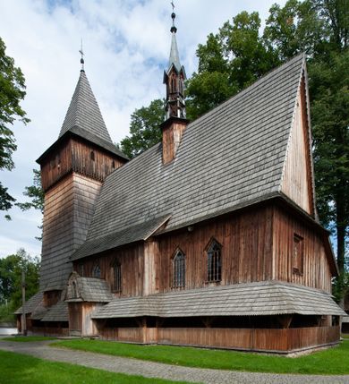 Zdjęcie nr 53: Kościół drewniany, orientowany, składający się z babińca, nawy głównej z przylegającą do niej od północy kaplicą boczną i kruchtą od południa oraz prezbiterium z zakrystią od północy. Prezbiterium i nawa w konstrukcji zrębowej, wieża, soboty i kruchta w słupowej. Kościół jednonawowy, nawa prawie kwadratowa, prezbiterium prostokątne, zamknięte prosto, oddzielone ścianą tęczową. Otwór tęczowy zamknięty łukiem dwuramiennym z biegnącą w poprzek grubą belką. Ściany gładkie, pokryte dekoracją malarską imitującą poziomo ułożone belki z uszczelniającymi je kiczkami, scenami figuralnymi, zacheuszkami i pasami ornamentalnymi. W nawie od zachodu chór muzyczny wsparty na dwóch, profilowanych słupach, z parapetem wybrzuszonym pośrodku; z dekoracją malarską o motywach geometryczno-roślinnych. Nawa i prezbiterium naryte pozornym sklepieniem z pułapami lekko zaokrąglonymi po bokach. Na całości przekrycia dekoracja malarska. W prezbiterium są to postacie aniołów wśród stylizowanych kwiatów na stropie adorujących monogram maryjny, nawę dekorują motywy roślinne. Anioły zdobią również sklepienie kaplicy. Strop babińca oraz fragmenty prezbiterium pokrywa oryginalna dekoracja patronowa złożona z motywów geometrycznych oraz figuralnych. W nawie głównej trzy okna, w prezbiterium i babińcu po dwa – od południa, kolejne do zakrystii; zamknięte łukiem dwuspadowym. Główne wejście od zachodu, drugie przez kruchtę od południa, trzecie do zakrystii. Portal południowy ostrołukowy, dekorowany ornamentem sznurowym, portal do zakrystii zamknięty łukiem w ośli grzbiet. Od zachodu kwadratowa wieża o pochyłych ścianach, z nadwieszoną izbicą, zwieńczona czteropołaciowym, ostrosłupowym hełmem gontowym. Izbica wieży oszalowana dookoła deskami zakończonymi u dołu ozdobnie wyrzynaną koronką. Od południa i wschodu soboty, czyli rodzaj podcieni,  w pozostałych miejscach gontowy kołnierz. Ściany świątyni oraz kaplicy są od zewnątrz szalowane deskami, wieża oraz przedsionek obite są gontem. Dachy i zadaszenia sobot pokryte gontem, kopuła kaplicy oraz wieżyczka na sygnaturkę pokryta blachą. 