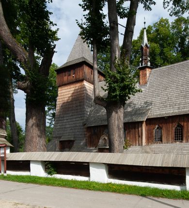 Zdjęcie nr 47: Kościół drewniany, orientowany, składający się z babińca, nawy głównej z przylegającą do niej od północy kaplicą boczną i kruchtą od południa oraz prezbiterium z zakrystią od północy. Prezbiterium i nawa w konstrukcji zrębowej, wieża, soboty i kruchta w słupowej. Kościół jednonawowy, nawa prawie kwadratowa, prezbiterium prostokątne, zamknięte prosto, oddzielone ścianą tęczową. Otwór tęczowy zamknięty łukiem dwuramiennym z biegnącą w poprzek grubą belką. Ściany gładkie, pokryte dekoracją malarską imitującą poziomo ułożone belki z uszczelniającymi je kiczkami, scenami figuralnymi, zacheuszkami i pasami ornamentalnymi. W nawie od zachodu chór muzyczny wsparty na dwóch, profilowanych słupach, z parapetem wybrzuszonym pośrodku; z dekoracją malarską o motywach geometryczno-roślinnych. Nawa i prezbiterium naryte pozornym sklepieniem z pułapami lekko zaokrąglonymi po bokach. Na całości przekrycia dekoracja malarska. W prezbiterium są to postacie aniołów wśród stylizowanych kwiatów na stropie adorujących monogram maryjny, nawę dekorują motywy roślinne. Anioły zdobią również sklepienie kaplicy. Strop babińca oraz fragmenty prezbiterium pokrywa oryginalna dekoracja patronowa złożona z motywów geometrycznych oraz figuralnych. W nawie głównej trzy okna, w prezbiterium i babińcu po dwa – od południa, kolejne do zakrystii; zamknięte łukiem dwuspadowym. Główne wejście od zachodu, drugie przez kruchtę od południa, trzecie do zakrystii. Portal południowy ostrołukowy, dekorowany ornamentem sznurowym, portal do zakrystii zamknięty łukiem w ośli grzbiet. Od zachodu kwadratowa wieża o pochyłych ścianach, z nadwieszoną izbicą, zwieńczona czteropołaciowym, ostrosłupowym hełmem gontowym. Izbica wieży oszalowana dookoła deskami zakończonymi u dołu ozdobnie wyrzynaną koronką. Od południa i wschodu soboty, czyli rodzaj podcieni,  w pozostałych miejscach gontowy kołnierz. Ściany świątyni oraz kaplicy są od zewnątrz szalowane deskami, wieża oraz przedsionek obite są gontem. Dachy i zadaszenia sobot pokryte gontem, kopuła kaplicy oraz wieżyczka na sygnaturkę pokryta blachą. 