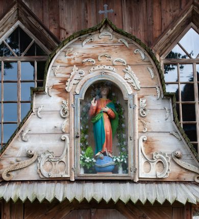 Zdjęcie nr 60: Kościół drewniany, orientowany, składający się z babińca, nawy głównej z przylegającą do niej od północy kaplicą boczną i kruchtą od południa oraz prezbiterium z zakrystią od północy. Prezbiterium i nawa w konstrukcji zrębowej, wieża, soboty i kruchta w słupowej. Kościół jednonawowy, nawa prawie kwadratowa, prezbiterium prostokątne, zamknięte prosto, oddzielone ścianą tęczową. Otwór tęczowy zamknięty łukiem dwuramiennym z biegnącą w poprzek grubą belką. Ściany gładkie, pokryte dekoracją malarską imitującą poziomo ułożone belki z uszczelniającymi je kiczkami, scenami figuralnymi, zacheuszkami i pasami ornamentalnymi. W nawie od zachodu chór muzyczny wsparty na dwóch, profilowanych słupach, z parapetem wybrzuszonym pośrodku; z dekoracją malarską o motywach geometryczno-roślinnych. Nawa i prezbiterium naryte pozornym sklepieniem z pułapami lekko zaokrąglonymi po bokach. Na całości przekrycia dekoracja malarska. W prezbiterium są to postacie aniołów wśród stylizowanych kwiatów na stropie adorujących monogram maryjny, nawę dekorują motywy roślinne. Anioły zdobią również sklepienie kaplicy. Strop babińca oraz fragmenty prezbiterium pokrywa oryginalna dekoracja patronowa złożona z motywów geometrycznych oraz figuralnych. W nawie głównej trzy okna, w prezbiterium i babińcu po dwa – od południa, kolejne do zakrystii; zamknięte łukiem dwuspadowym. Główne wejście od zachodu, drugie przez kruchtę od południa, trzecie do zakrystii. Portal południowy ostrołukowy, dekorowany ornamentem sznurowym, portal do zakrystii zamknięty łukiem w ośli grzbiet. Od zachodu kwadratowa wieża o pochyłych ścianach, z nadwieszoną izbicą, zwieńczona czteropołaciowym, ostrosłupowym hełmem gontowym. Izbica wieży oszalowana dookoła deskami zakończonymi u dołu ozdobnie wyrzynaną koronką. Od południa i wschodu soboty, czyli rodzaj podcieni,  w pozostałych miejscach gontowy kołnierz. Ściany świątyni oraz kaplicy są od zewnątrz szalowane deskami, wieża oraz przedsionek obite są gontem. Dachy i zadaszenia sobot pokryte gontem, kopuła kaplicy oraz wieżyczka na sygnaturkę pokryta blachą. 