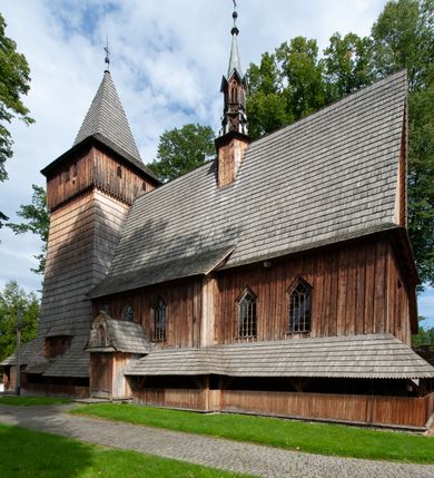 Zdjęcie nr 56: Kościół drewniany, orientowany, składający się z babińca, nawy głównej z przylegającą do niej od północy kaplicą boczną i kruchtą od południa oraz prezbiterium z zakrystią od północy. Prezbiterium i nawa w konstrukcji zrębowej, wieża, soboty i kruchta w słupowej. Kościół jednonawowy, nawa prawie kwadratowa, prezbiterium prostokątne, zamknięte prosto, oddzielone ścianą tęczową. Otwór tęczowy zamknięty łukiem dwuramiennym z biegnącą w poprzek grubą belką. Ściany gładkie, pokryte dekoracją malarską imitującą poziomo ułożone belki z uszczelniającymi je kiczkami, scenami figuralnymi, zacheuszkami i pasami ornamentalnymi. W nawie od zachodu chór muzyczny wsparty na dwóch, profilowanych słupach, z parapetem wybrzuszonym pośrodku; z dekoracją malarską o motywach geometryczno-roślinnych. Nawa i prezbiterium naryte pozornym sklepieniem z pułapami lekko zaokrąglonymi po bokach. Na całości przekrycia dekoracja malarska. W prezbiterium są to postacie aniołów wśród stylizowanych kwiatów na stropie adorujących monogram maryjny, nawę dekorują motywy roślinne. Anioły zdobią również sklepienie kaplicy. Strop babińca oraz fragmenty prezbiterium pokrywa oryginalna dekoracja patronowa złożona z motywów geometrycznych oraz figuralnych. W nawie głównej trzy okna, w prezbiterium i babińcu po dwa – od południa, kolejne do zakrystii; zamknięte łukiem dwuspadowym. Główne wejście od zachodu, drugie przez kruchtę od południa, trzecie do zakrystii. Portal południowy ostrołukowy, dekorowany ornamentem sznurowym, portal do zakrystii zamknięty łukiem w ośli grzbiet. Od zachodu kwadratowa wieża o pochyłych ścianach, z nadwieszoną izbicą, zwieńczona czteropołaciowym, ostrosłupowym hełmem gontowym. Izbica wieży oszalowana dookoła deskami zakończonymi u dołu ozdobnie wyrzynaną koronką. Od południa i wschodu soboty, czyli rodzaj podcieni,  w pozostałych miejscach gontowy kołnierz. Ściany świątyni oraz kaplicy są od zewnątrz szalowane deskami, wieża oraz przedsionek obite są gontem. Dachy i zadaszenia sobot pokryte gontem, kopuła kaplicy oraz wieżyczka na sygnaturkę pokryta blachą. 
