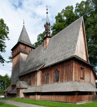 Zdjęcie nr 55: Kościół drewniany, orientowany, składający się z babińca, nawy głównej z przylegającą do niej od północy kaplicą boczną i kruchtą od południa oraz prezbiterium z zakrystią od północy. Prezbiterium i nawa w konstrukcji zrębowej, wieża, soboty i kruchta w słupowej. Kościół jednonawowy, nawa prawie kwadratowa, prezbiterium prostokątne, zamknięte prosto, oddzielone ścianą tęczową. Otwór tęczowy zamknięty łukiem dwuramiennym z biegnącą w poprzek grubą belką. Ściany gładkie, pokryte dekoracją malarską imitującą poziomo ułożone belki z uszczelniającymi je kiczkami, scenami figuralnymi, zacheuszkami i pasami ornamentalnymi. W nawie od zachodu chór muzyczny wsparty na dwóch, profilowanych słupach, z parapetem wybrzuszonym pośrodku; z dekoracją malarską o motywach geometryczno-roślinnych. Nawa i prezbiterium naryte pozornym sklepieniem z pułapami lekko zaokrąglonymi po bokach. Na całości przekrycia dekoracja malarska. W prezbiterium są to postacie aniołów wśród stylizowanych kwiatów na stropie adorujących monogram maryjny, nawę dekorują motywy roślinne. Anioły zdobią również sklepienie kaplicy. Strop babińca oraz fragmenty prezbiterium pokrywa oryginalna dekoracja patronowa złożona z motywów geometrycznych oraz figuralnych. W nawie głównej trzy okna, w prezbiterium i babińcu po dwa – od południa, kolejne do zakrystii; zamknięte łukiem dwuspadowym. Główne wejście od zachodu, drugie przez kruchtę od południa, trzecie do zakrystii. Portal południowy ostrołukowy, dekorowany ornamentem sznurowym, portal do zakrystii zamknięty łukiem w ośli grzbiet. Od zachodu kwadratowa wieża o pochyłych ścianach, z nadwieszoną izbicą, zwieńczona czteropołaciowym, ostrosłupowym hełmem gontowym. Izbica wieży oszalowana dookoła deskami zakończonymi u dołu ozdobnie wyrzynaną koronką. Od południa i wschodu soboty, czyli rodzaj podcieni,  w pozostałych miejscach gontowy kołnierz. Ściany świątyni oraz kaplicy są od zewnątrz szalowane deskami, wieża oraz przedsionek obite są gontem. Dachy i zadaszenia sobot pokryte gontem, kopuła kaplicy oraz wieżyczka na sygnaturkę pokryta blachą. 