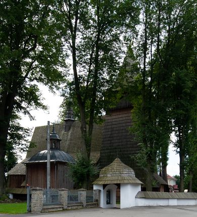 Zdjęcie nr 45: Kościół drewniany, orientowany, składający się z babińca, nawy głównej z przylegającą do niej od północy kaplicą boczną i kruchtą od południa oraz prezbiterium z zakrystią od północy. Prezbiterium i nawa w konstrukcji zrębowej, wieża, soboty i kruchta w słupowej. Kościół jednonawowy, nawa prawie kwadratowa, prezbiterium prostokątne, zamknięte prosto, oddzielone ścianą tęczową. Otwór tęczowy zamknięty łukiem dwuramiennym z biegnącą w poprzek grubą belką. Ściany gładkie, pokryte dekoracją malarską imitującą poziomo ułożone belki z uszczelniającymi je kiczkami, scenami figuralnymi, zacheuszkami i pasami ornamentalnymi. W nawie od zachodu chór muzyczny wsparty na dwóch, profilowanych słupach, z parapetem wybrzuszonym pośrodku; z dekoracją malarską o motywach geometryczno-roślinnych. Nawa i prezbiterium naryte pozornym sklepieniem z pułapami lekko zaokrąglonymi po bokach. Na całości przekrycia dekoracja malarska. W prezbiterium są to postacie aniołów wśród stylizowanych kwiatów na stropie adorujących monogram maryjny, nawę dekorują motywy roślinne. Anioły zdobią również sklepienie kaplicy. Strop babińca oraz fragmenty prezbiterium pokrywa oryginalna dekoracja patronowa złożona z motywów geometrycznych oraz figuralnych. W nawie głównej trzy okna, w prezbiterium i babińcu po dwa – od południa, kolejne do zakrystii; zamknięte łukiem dwuspadowym. Główne wejście od zachodu, drugie przez kruchtę od południa, trzecie do zakrystii. Portal południowy ostrołukowy, dekorowany ornamentem sznurowym, portal do zakrystii zamknięty łukiem w ośli grzbiet. Od zachodu kwadratowa wieża o pochyłych ścianach, z nadwieszoną izbicą, zwieńczona czteropołaciowym, ostrosłupowym hełmem gontowym. Izbica wieży oszalowana dookoła deskami zakończonymi u dołu ozdobnie wyrzynaną koronką. Od południa i wschodu soboty, czyli rodzaj podcieni,  w pozostałych miejscach gontowy kołnierz. Ściany świątyni oraz kaplicy są od zewnątrz szalowane deskami, wieża oraz przedsionek obite są gontem. Dachy i zadaszenia sobot pokryte gontem, kopuła kaplicy oraz wieżyczka na sygnaturkę pokryta blachą. 