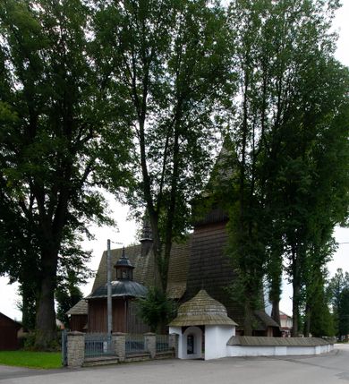 Zdjęcie nr 44: Kościół drewniany, orientowany, składający się z babińca, nawy głównej z przylegającą do niej od północy kaplicą boczną i kruchtą od południa oraz prezbiterium z zakrystią od północy. Prezbiterium i nawa w konstrukcji zrębowej, wieża, soboty i kruchta w słupowej. Kościół jednonawowy, nawa prawie kwadratowa, prezbiterium prostokątne, zamknięte prosto, oddzielone ścianą tęczową. Otwór tęczowy zamknięty łukiem dwuramiennym z biegnącą w poprzek grubą belką. Ściany gładkie, pokryte dekoracją malarską imitującą poziomo ułożone belki z uszczelniającymi je kiczkami, scenami figuralnymi, zacheuszkami i pasami ornamentalnymi. W nawie od zachodu chór muzyczny wsparty na dwóch, profilowanych słupach, z parapetem wybrzuszonym pośrodku; z dekoracją malarską o motywach geometryczno-roślinnych. Nawa i prezbiterium naryte pozornym sklepieniem z pułapami lekko zaokrąglonymi po bokach. Na całości przekrycia dekoracja malarska. W prezbiterium są to postacie aniołów wśród stylizowanych kwiatów na stropie adorujących monogram maryjny, nawę dekorują motywy roślinne. Anioły zdobią również sklepienie kaplicy. Strop babińca oraz fragmenty prezbiterium pokrywa oryginalna dekoracja patronowa złożona z motywów geometrycznych oraz figuralnych. W nawie głównej trzy okna, w prezbiterium i babińcu po dwa – od południa, kolejne do zakrystii; zamknięte łukiem dwuspadowym. Główne wejście od zachodu, drugie przez kruchtę od południa, trzecie do zakrystii. Portal południowy ostrołukowy, dekorowany ornamentem sznurowym, portal do zakrystii zamknięty łukiem w ośli grzbiet. Od zachodu kwadratowa wieża o pochyłych ścianach, z nadwieszoną izbicą, zwieńczona czteropołaciowym, ostrosłupowym hełmem gontowym. Izbica wieży oszalowana dookoła deskami zakończonymi u dołu ozdobnie wyrzynaną koronką. Od południa i wschodu soboty, czyli rodzaj podcieni,  w pozostałych miejscach gontowy kołnierz. Ściany świątyni oraz kaplicy są od zewnątrz szalowane deskami, wieża oraz przedsionek obite są gontem. Dachy i zadaszenia sobot pokryte gontem, kopuła kaplicy oraz wieżyczka na sygnaturkę pokryta blachą. 