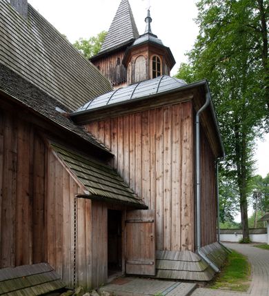 Zdjęcie nr 64: Kościół drewniany, orientowany, składający się z babińca, nawy głównej z przylegającą do niej od północy kaplicą boczną i kruchtą od południa oraz prezbiterium z zakrystią od północy. Prezbiterium i nawa w konstrukcji zrębowej, wieża, soboty i kruchta w słupowej. Kościół jednonawowy, nawa prawie kwadratowa, prezbiterium prostokątne, zamknięte prosto, oddzielone ścianą tęczową. Otwór tęczowy zamknięty łukiem dwuramiennym z biegnącą w poprzek grubą belką. Ściany gładkie, pokryte dekoracją malarską imitującą poziomo ułożone belki z uszczelniającymi je kiczkami, scenami figuralnymi, zacheuszkami i pasami ornamentalnymi. W nawie od zachodu chór muzyczny wsparty na dwóch, profilowanych słupach, z parapetem wybrzuszonym pośrodku; z dekoracją malarską o motywach geometryczno-roślinnych. Nawa i prezbiterium naryte pozornym sklepieniem z pułapami lekko zaokrąglonymi po bokach. Na całości przekrycia dekoracja malarska. W prezbiterium są to postacie aniołów wśród stylizowanych kwiatów na stropie adorujących monogram maryjny, nawę dekorują motywy roślinne. Anioły zdobią również sklepienie kaplicy. Strop babińca oraz fragmenty prezbiterium pokrywa oryginalna dekoracja patronowa złożona z motywów geometrycznych oraz figuralnych. W nawie głównej trzy okna, w prezbiterium i babińcu po dwa – od południa, kolejne do zakrystii; zamknięte łukiem dwuspadowym. Główne wejście od zachodu, drugie przez kruchtę od południa, trzecie do zakrystii. Portal południowy ostrołukowy, dekorowany ornamentem sznurowym, portal do zakrystii zamknięty łukiem w ośli grzbiet. Od zachodu kwadratowa wieża o pochyłych ścianach, z nadwieszoną izbicą, zwieńczona czteropołaciowym, ostrosłupowym hełmem gontowym. Izbica wieży oszalowana dookoła deskami zakończonymi u dołu ozdobnie wyrzynaną koronką. Od południa i wschodu soboty, czyli rodzaj podcieni,  w pozostałych miejscach gontowy kołnierz. Ściany świątyni oraz kaplicy są od zewnątrz szalowane deskami, wieża oraz przedsionek obite są gontem. Dachy i zadaszenia sobot pokryte gontem, kopuła kaplicy oraz wieżyczka na sygnaturkę pokryta blachą. 