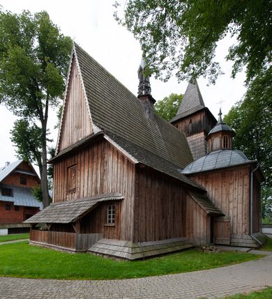 Zdjęcie nr 62: Kościół drewniany, orientowany, składający się z babińca, nawy głównej z przylegającą do niej od północy kaplicą boczną i kruchtą od południa oraz prezbiterium z zakrystią od północy. Prezbiterium i nawa w konstrukcji zrębowej, wieża, soboty i kruchta w słupowej. Kościół jednonawowy, nawa prawie kwadratowa, prezbiterium prostokątne, zamknięte prosto, oddzielone ścianą tęczową. Otwór tęczowy zamknięty łukiem dwuramiennym z biegnącą w poprzek grubą belką. Ściany gładkie, pokryte dekoracją malarską imitującą poziomo ułożone belki z uszczelniającymi je kiczkami, scenami figuralnymi, zacheuszkami i pasami ornamentalnymi. W nawie od zachodu chór muzyczny wsparty na dwóch, profilowanych słupach, z parapetem wybrzuszonym pośrodku; z dekoracją malarską o motywach geometryczno-roślinnych. Nawa i prezbiterium naryte pozornym sklepieniem z pułapami lekko zaokrąglonymi po bokach. Na całości przekrycia dekoracja malarska. W prezbiterium są to postacie aniołów wśród stylizowanych kwiatów na stropie adorujących monogram maryjny, nawę dekorują motywy roślinne. Anioły zdobią również sklepienie kaplicy. Strop babińca oraz fragmenty prezbiterium pokrywa oryginalna dekoracja patronowa złożona z motywów geometrycznych oraz figuralnych. W nawie głównej trzy okna, w prezbiterium i babińcu po dwa – od południa, kolejne do zakrystii; zamknięte łukiem dwuspadowym. Główne wejście od zachodu, drugie przez kruchtę od południa, trzecie do zakrystii. Portal południowy ostrołukowy, dekorowany ornamentem sznurowym, portal do zakrystii zamknięty łukiem w ośli grzbiet. Od zachodu kwadratowa wieża o pochyłych ścianach, z nadwieszoną izbicą, zwieńczona czteropołaciowym, ostrosłupowym hełmem gontowym. Izbica wieży oszalowana dookoła deskami zakończonymi u dołu ozdobnie wyrzynaną koronką. Od południa i wschodu soboty, czyli rodzaj podcieni,  w pozostałych miejscach gontowy kołnierz. Ściany świątyni oraz kaplicy są od zewnątrz szalowane deskami, wieża oraz przedsionek obite są gontem. Dachy i zadaszenia sobot pokryte gontem, kopuła kaplicy oraz wieżyczka na sygnaturkę pokryta blachą. 