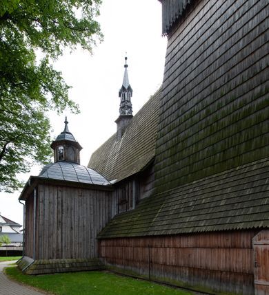 Zdjęcie nr 65: Kościół drewniany, orientowany, składający się z babińca, nawy głównej z przylegającą do niej od północy kaplicą boczną i kruchtą od południa oraz prezbiterium z zakrystią od północy. Prezbiterium i nawa w konstrukcji zrębowej, wieża, soboty i kruchta w słupowej. Kościół jednonawowy, nawa prawie kwadratowa, prezbiterium prostokątne, zamknięte prosto, oddzielone ścianą tęczową. Otwór tęczowy zamknięty łukiem dwuramiennym z biegnącą w poprzek grubą belką. Ściany gładkie, pokryte dekoracją malarską imitującą poziomo ułożone belki z uszczelniającymi je kiczkami, scenami figuralnymi, zacheuszkami i pasami ornamentalnymi. W nawie od zachodu chór muzyczny wsparty na dwóch, profilowanych słupach, z parapetem wybrzuszonym pośrodku; z dekoracją malarską o motywach geometryczno-roślinnych. Nawa i prezbiterium naryte pozornym sklepieniem z pułapami lekko zaokrąglonymi po bokach. Na całości przekrycia dekoracja malarska. W prezbiterium są to postacie aniołów wśród stylizowanych kwiatów na stropie adorujących monogram maryjny, nawę dekorują motywy roślinne. Anioły zdobią również sklepienie kaplicy. Strop babińca oraz fragmenty prezbiterium pokrywa oryginalna dekoracja patronowa złożona z motywów geometrycznych oraz figuralnych. W nawie głównej trzy okna, w prezbiterium i babińcu po dwa – od południa, kolejne do zakrystii; zamknięte łukiem dwuspadowym. Główne wejście od zachodu, drugie przez kruchtę od południa, trzecie do zakrystii. Portal południowy ostrołukowy, dekorowany ornamentem sznurowym, portal do zakrystii zamknięty łukiem w ośli grzbiet. Od zachodu kwadratowa wieża o pochyłych ścianach, z nadwieszoną izbicą, zwieńczona czteropołaciowym, ostrosłupowym hełmem gontowym. Izbica wieży oszalowana dookoła deskami zakończonymi u dołu ozdobnie wyrzynaną koronką. Od południa i wschodu soboty, czyli rodzaj podcieni,  w pozostałych miejscach gontowy kołnierz. Ściany świątyni oraz kaplicy są od zewnątrz szalowane deskami, wieża oraz przedsionek obite są gontem. Dachy i zadaszenia sobot pokryte gontem, kopuła kaplicy oraz wieżyczka na sygnaturkę pokryta blachą. 