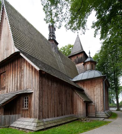 Zdjęcie nr 63: Kościół drewniany, orientowany, składający się z babińca, nawy głównej z przylegającą do niej od północy kaplicą boczną i kruchtą od południa oraz prezbiterium z zakrystią od północy. Prezbiterium i nawa w konstrukcji zrębowej, wieża, soboty i kruchta w słupowej. Kościół jednonawowy, nawa prawie kwadratowa, prezbiterium prostokątne, zamknięte prosto, oddzielone ścianą tęczową. Otwór tęczowy zamknięty łukiem dwuramiennym z biegnącą w poprzek grubą belką. Ściany gładkie, pokryte dekoracją malarską imitującą poziomo ułożone belki z uszczelniającymi je kiczkami, scenami figuralnymi, zacheuszkami i pasami ornamentalnymi. W nawie od zachodu chór muzyczny wsparty na dwóch, profilowanych słupach, z parapetem wybrzuszonym pośrodku; z dekoracją malarską o motywach geometryczno-roślinnych. Nawa i prezbiterium naryte pozornym sklepieniem z pułapami lekko zaokrąglonymi po bokach. Na całości przekrycia dekoracja malarska. W prezbiterium są to postacie aniołów wśród stylizowanych kwiatów na stropie adorujących monogram maryjny, nawę dekorują motywy roślinne. Anioły zdobią również sklepienie kaplicy. Strop babińca oraz fragmenty prezbiterium pokrywa oryginalna dekoracja patronowa złożona z motywów geometrycznych oraz figuralnych. W nawie głównej trzy okna, w prezbiterium i babińcu po dwa – od południa, kolejne do zakrystii; zamknięte łukiem dwuspadowym. Główne wejście od zachodu, drugie przez kruchtę od południa, trzecie do zakrystii. Portal południowy ostrołukowy, dekorowany ornamentem sznurowym, portal do zakrystii zamknięty łukiem w ośli grzbiet. Od zachodu kwadratowa wieża o pochyłych ścianach, z nadwieszoną izbicą, zwieńczona czteropołaciowym, ostrosłupowym hełmem gontowym. Izbica wieży oszalowana dookoła deskami zakończonymi u dołu ozdobnie wyrzynaną koronką. Od południa i wschodu soboty, czyli rodzaj podcieni,  w pozostałych miejscach gontowy kołnierz. Ściany świątyni oraz kaplicy są od zewnątrz szalowane deskami, wieża oraz przedsionek obite są gontem. Dachy i zadaszenia sobot pokryte gontem, kopuła kaplicy oraz wieżyczka na sygnaturkę pokryta blachą. 