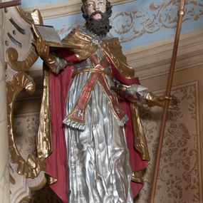 Zdjęcie nr 1: Rzeźba ustawiona na niskim, prostopadłościennym cokole przedstawiająca św. Wojciecha w postawie stojącej w delikatnym kontrapoście, z pastorałem w lewej dłoni, z otwartą księgą w prawej, z głową skierowaną ku górze, lewy bok przebity włócznią. Twarz podłużna, okolona długą, ciemnobrązową, bujną brodą, oczy duże, nos długi i szeroki, włosy średniej długości, kręcone. Święty jest ubrany w albę z długimi rękawami, złoconą kapę z czerwoną podszewką spiętą na piersi klamrą, spod kapy widoczna czerwona stuła, na stopach złocone buty, a na głowie infuła, na dłoniach złote rękawice. Polichromia w odsłoniętych partiach ciała naturalistyczna. 