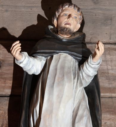 Zdjęcie nr 1: Figura nieokreślonego świętego dominikańskiego, pełnoplastyczna, przyścienna, wydrążona i spłaszczona od tyłu, w ujęciu frontalnym, umieszczona na cokoliku. Sylwetka świętego lekko wygięta w kształcie litery S, w kontrapoście, z rękoma ugiętymi w łokciach, odwiedzionymi od ciała na boki i wzniesionymi ku górze na wysokość ramion. Twarz lekko wydłużona z zarysowanymi kośćmi policzkowymi, jasna karnacja, niskie czoło, spiczasty nos i wąskie, lekko rozchylone usta. Małe, głęboko osadzone oczy, wydatne uszy. Włosy i broda koloru brązowego. Na głowie tonsura. Święty ubrany w biały habit dominikański ze szkaplerzem oraz czarną kapę z kapturem. Spod habitu wyłaniają się brązowe buty. Szaty podkreślające anatomię. Fałdy szat, głęboko wycięte na habicie, a płytko na szkaplerzu, dynamizują postać. Układają się u dołu w formy trójkątne.