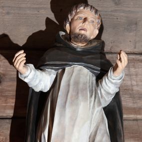 Zdjęcie nr 1: Figura nieokreślonego świętego dominikańskiego, pełnoplastyczna, przyścienna, wydrążona i spłaszczona od tyłu, w ujęciu frontalnym, umieszczona na cokoliku. Sylwetka świętego lekko wygięta w kształcie litery S, w kontrapoście, z rękoma ugiętymi w łokciach, odwiedzionymi od ciała na boki i wzniesionymi ku górze na wysokość ramion. Twarz lekko wydłużona z zarysowanymi kośćmi policzkowymi, jasna karnacja, niskie czoło, spiczasty nos i wąskie, lekko rozchylone usta. Małe, głęboko osadzone oczy, wydatne uszy. Włosy i broda koloru brązowego. Na głowie tonsura. Święty ubrany w biały habit dominikański ze szkaplerzem oraz czarną kapę z kapturem. Spod habitu wyłaniają się brązowe buty. Szaty podkreślające anatomię. Fałdy szat, głęboko wycięte na habicie, a płytko na szkaplerzu, dynamizują postać. Układają się u dołu w formy trójkątne.