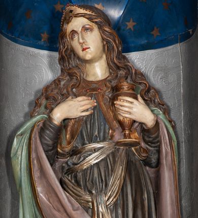 Zdjęcie nr 1: Rzeźba pełnoplastyczna, całopostaciowa, przyścienna, drążona z tyłu. Św. Maria Magdalena ukazana została w pozycji stojącej, z lekko zgiętą w kolanie, wysuniętą do przodu prawą nogą. Prawa ręka zgięta w łokciu spoczywa na piersi, w lewej dłoni święta trzyma kielich. Jej głowa jest lekko zwrócona w prawą stronę, a wzrok skierowany ku górze. Długie pofalowane włosy opadają na ramiona. Na głowie św. Maria Magdalena nałożoną ma złoty diadem. Postać ubrana jest w długą szatę, z długimi rękawami, w kolorze szaroniebieskim, zdobioną złotą lamówką. Biodra świętej przepasane są szarfą w kolorze złotym. Na ramiona narzucony ma błękitny płaszcz, spięty pod szyją klamrą. Poły płaszcza przytrzymywane są przez św. Marie Magdalenę na przedramionach i spływają po zewnętrznych ich stronach. Prawa poła płaszcza zakrywa prawe kolano, wywijając się i ukazując różowe podbicie. U stóp Świętej, po lewej stronie znajduje się czaszka.