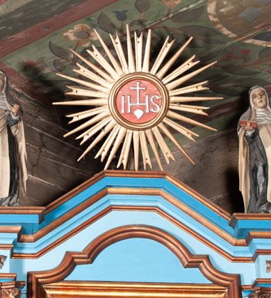 Zdjęcie nr 2: Nastawa ołtarzowa architektoniczna, jednoosiowa, jednokondygnacyjna. Ołtarz drewniany, sarkofagowy, w formie odwróconego trapezu. Mensa drewniana, silnie wysunięta. Antependium w formie odwróconego trapezu, na cokole, w polu romb ujęty ramą z rozetą. Ołtarz flankują romby ujęte ramą.
Retabulum na dwustrefowym cokole, zryzalitowanym na skraju; w konsole dolnej strefy dźwigaja cokoły górnej. Ryzality podtrzymują parę półfilarów o stylizowanych kapitelach kompozytowych i kanelowanych trzonach. Podpory dźwigają pełne belkowanie, wyłamane na ich osi, z silnie wysuniętym, bogato profilowanym gzymsem; nad polem środkowym niepełne, przyjmujące kształt trzech boków z ośmioboku; Na gzymsie, na osi podpór figury dwóch karmelitańskich świętych. W polu środkowym nisza w formie stojącego prostokąta zamkniętego łukiem nadwieszonym z uskokiem, ujęta profilowaną ramą; wewnątrz profilowana rama w formie stojącego prostokąta, w której znajduje się obraz Najświętsze Serce Jezusa w ramie. Na belkowaniu, nad polem środkowym medalion z monogramem IHS w glorii promienistej, którego podstawę stanowi fragment rocaille.
W górnej strefie cokołu dwie płyciny w formie leżącego prostokąta z rozetami; cokoły kolumn zrobione z trzech stron płycinami z malowaną dekoracją roślinną. W partii fryzu na osi podpór rozety. Rama niszy zwieńczona elementem rocaille. Retabulum ujęte uszami utworzonymi z suchego akantu.
Struktura polichromowana w kolorze jasnoniebieskim, profile na niebiesko; konsole i trzony marmoryzowane; ornamentyka, ramy, profile i gloria złocone.
