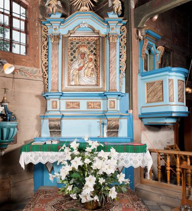 Zdjęcie nr 2: Nastawa ołtarzowa architektoniczna, jednoosiowa, jednokondygnacyjna, ze zwieńczeniem. Ołtarz drewniany, sarkofagowy, zwężający się ku dołowi, na cokole. Mensa drewniana, silnie wysunięta.  Antependium w kształcie trapezu, w polu romb ujęty obramieniem, z rozetą, flankowany dwoma rombami. Retabulum na dwustrefowym cokole, flankowane para półfilarów o stylizowanych kapitelach kompozytowych i kanelowanych trzonach, umieszczonych na cokołach nadwieszonych na konsolach. Podpory dźwigają pełne belkowanie, występujące na ich osi, z silnie wysuniętym, bogato profilowanym gzymsem, nad polem środkowym wyłamanym trójbocznie. W zwieńczeniu w osiach podpór odcinki przerwanego przyczółka, na których siedzą dwa uskrzydlone putta. W polu środkowym nisza w kształcie stojącego prostokąta zamkniętego łukiem nadwieszonym z uskokiem, ujęta profilowaną ramą; wewnątrz profilowana rama w kształcie stojącego prostokąta, w której znajduje się obraz Matki Boskiej z Dzieciątkiem w ramie dekorowanej płaskorzeźbionymi motywami liści akantu. W zwieńczeniu, nad polem środkowym medalion z ze splecionymi literami imienia Marya w glorii promienistej, u dołu motyw rocaille’u. W górnej strefie cokołu dwie płyciny w formie leżącego prostokąta z rozetami; cokoły kolumn zdobione z trzech stron płycinami z malowaną dekoracją roślinną. W partii fryzu na osi podpór rozety. Rama niszy zwieńczona motywem rocaille’u. Retabulum ujęte uszami utworzonymi z motywów stylizowanych liści akantu i elementów małżowiny. Struktura polichromowana w kolorze jasnoniebieskim, profile na niebiesko; konsole i trzony marmoryzowane; ornamentyka, ramy, profile i gloria złocone.