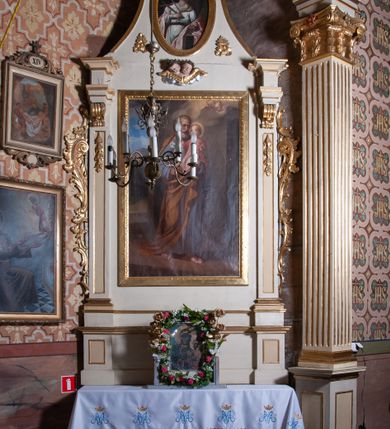 Zdjęcie nr 5: Ołtarz architektoniczny, jednoosiowy, jednokondygnacyjny ze zwieńczeniem, z uszami. Retabulum ustawione na prostopadłościennej mensie ołtarzowej z antepedium, zdobionym czterema prostokątnymi płycinami oddzielonymi kanelowanymi lizenami. Tabernakulum w kształcie prostopadłościennej skrzynki na wysokim cokole, zamkniętej od góry płaskim daszkiem, na którym miejsce na prosty tron do wystawiania monstrancji. Przednia ścianka tabernakulum zamknięta od góry gzymsem w formie łuku nadwieszonego. Drzwiczki w formie stojącego prostokąta zamkniętego łukiem półkolistym. W polu, obwiedzionym ramą o analogicznym kształcie płaskorzeźbiony krzyż z podstawą. Ustawiona na mensie nastawa wsparta na cokole z wyodrębnionymi po bokach dwoma postumentami, wyżej ujęta parą pilastrów, o trzonach ozdobionych płycinami i u góry zwisami ze stylizowanych kampanul, z nałożonymi u szczytu konsolkami pełniącymi funkcję kapiteli, które wspierają odcinki przerwanego belkowania. W polu głównym obraz św. Józefa z Dzieciątkiem Jezus w kształcie pionowego prostokąta. Zwieńczenie  w ogólnym zarysie w kształcie trapezu, o bokach zaakcentowanych wklęsłymi łukami odcinkowymi, zamknięte od góry dwiema wolutkami, spiętymi u szczytu muszlą palmetową i ozdobionymi wicią z różami. W polu owalny obraz z przedstawieniem św. Wiktorii. Poniżej dwie uskrzydlone główki aniołków. Uszy z wici i motywów ornamentu rocaille&#039;owego. Struktura ołtarza w kolorze białym, detale i ornamentyka złocone.