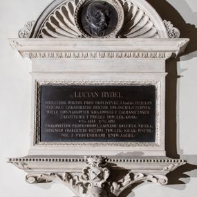 Zdjęcie nr 1: Epitafium w formie leżącego prostokąta, wsparte na szerokim, wydatnym gzymsie z perełkowaniem, z narożami ujętymi liśćmi, z konsolą z wolutami i liśćmi oraz uskrzydloną główką anioła oraz owalnym herbem Uniwersytetu Jagiellońskiego w ozdobnym kartuszu, zwieńczonym koroną. Tablicę wieńczy gzyms z motywem wolich oczu, na którym osadzone jest półkoliste zwieńczenie z akroterionami, wypełnione muszlą palmetową i okrągłym medalionem obwiedzionym motywem wolich oczu z płaskorzeźbionym wizerunkiem zmarłego. Postać ukazana jest w popiersiu, zwrócona w lewo, o twarzy wyrazistej, z wąsami i wysokim czołem z zakolami. W marmurowej tablicy inskrypcja: „LUCYAN RYDEL / MED(YCYNY) I CHIR(URGII): DOKTOR PROF(ESOR) OKULISTYKI, 3 krotnie DZIEKAN / WYDZIAŁU LEKARSKIEGO, REKTOR UNI(WERSYTETU) JAGIEL(LOŃSKIEGO) CZŁONEK / WIELU TOW(ARZYSTW) NAUKOWYCH KRAJOWYCH I ZAGRANICZNYCH / ZAŁOŻYCIEL I PREZES TOW(ARZYSTWA) LEK(ARSKIEGO) KRAK(OWSKIEGO) / *17/10 1833 †27/4 1895 / ZNAKOMITEMU PROFESOROWI, ZACNEMU KOLEDZE, NIESKA/ZITELNEGO CHARAKTERU MĘŻOWI, TOW(ARZYSTWO) LEK(ARSKIE) KRAK(OWSKIE) WSPOL/NIE Z PROFESORAMI UNIW(ERSYTETU) JAGIEL(LOŃSKIEGO)”. Na medalionie sygnatura: „Z[ygmunt] Langman / Kraków”.