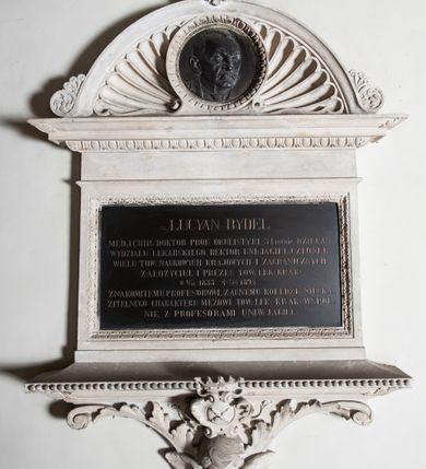 Zdjęcie nr 3: Epitafium w formie leżącego prostokąta, wsparte na szerokim, wydatnym gzymsie z perełkowaniem, z narożami ujętymi liśćmi, z konsolą z wolutami i liśćmi oraz uskrzydloną główką anioła oraz owalnym herbem Uniwersytetu Jagiellońskiego w ozdobnym kartuszu, zwieńczonym koroną. Tablicę wieńczy gzyms z motywem wolich oczu, na którym osadzone jest półkoliste zwieńczenie z akroterionami, wypełnione muszlą palmetową i okrągłym medalionem obwiedzionym motywem wolich oczu z płaskorzeźbionym wizerunkiem zmarłego. Postać ukazana jest w popiersiu, zwrócona w lewo, o twarzy wyrazistej, z wąsami i wysokim czołem z zakolami. W marmurowej tablicy inskrypcja: „LUCYAN RYDEL / MED(YCYNY) I CHIR(URGII): DOKTOR PROF(ESOR) OKULISTYKI, 3 krotnie DZIEKAN / WYDZIAŁU LEKARSKIEGO, REKTOR UNI(WERSYTETU) JAGIEL(LOŃSKIEGO) CZŁONEK / WIELU TOW(ARZYSTW) NAUKOWYCH KRAJOWYCH I ZAGRANICZNYCH / ZAŁOŻYCIEL I PREZES TOW(ARZYSTWA) LEK(ARSKIEGO) KRAK(OWSKIEGO) / *17/10 1833 †27/4 1895 / ZNAKOMITEMU PROFESOROWI, ZACNEMU KOLEDZE, NIESKA/ZITELNEGO CHARAKTERU MĘŻOWI, TOW(ARZYSTWO) LEK(ARSKIE) KRAK(OWSKIE) WSPOL/NIE Z PROFESORAMI UNIW(ERSYTETU) JAGIEL(LOŃSKIEGO)”. Na medalionie sygnatura: „Z[ygmunt] Langman / Kraków”.