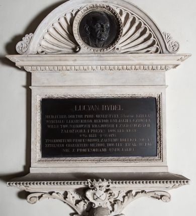 Zdjęcie nr 2: Epitafium w formie leżącego prostokąta, wsparte na szerokim, wydatnym gzymsie z perełkowaniem, z narożami ujętymi liśćmi, z konsolą z wolutami i liśćmi oraz uskrzydloną główką anioła oraz owalnym herbem Uniwersytetu Jagiellońskiego w ozdobnym kartuszu, zwieńczonym koroną. Tablicę wieńczy gzyms z motywem wolich oczu, na którym osadzone jest półkoliste zwieńczenie z akroterionami, wypełnione muszlą palmetową i okrągłym medalionem obwiedzionym motywem wolich oczu z płaskorzeźbionym wizerunkiem zmarłego. Postać ukazana jest w popiersiu, zwrócona w lewo, o twarzy wyrazistej, z wąsami i wysokim czołem z zakolami. W marmurowej tablicy inskrypcja: „LUCYAN RYDEL / MED(YCYNY) I CHIR(URGII): DOKTOR PROF(ESOR) OKULISTYKI, 3 krotnie DZIEKAN / WYDZIAŁU LEKARSKIEGO, REKTOR UNI(WERSYTETU) JAGIEL(LOŃSKIEGO) CZŁONEK / WIELU TOW(ARZYSTW) NAUKOWYCH KRAJOWYCH I ZAGRANICZNYCH / ZAŁOŻYCIEL I PREZES TOW(ARZYSTWA) LEK(ARSKIEGO) KRAK(OWSKIEGO) / *17/10 1833 †27/4 1895 / ZNAKOMITEMU PROFESOROWI, ZACNEMU KOLEDZE, NIESKA/ZITELNEGO CHARAKTERU MĘŻOWI, TOW(ARZYSTWO) LEK(ARSKIE) KRAK(OWSKIE) WSPOL/NIE Z PROFESORAMI UNIW(ERSYTETU) JAGIEL(LOŃSKIEGO)”. Na medalionie sygnatura: „Z[ygmunt] Langman / Kraków”.