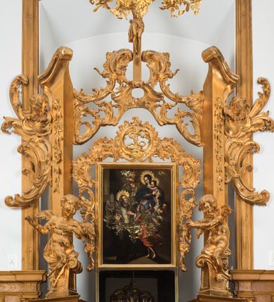 Zdjęcie nr 4: Na brązowym tle, z kolumną w lewej części kompozycji i lekko rozjaśnionymi obłokami, ukazany św. Jan Kanty klęczący przed siedzącą w chmurach Matką Boską z Dzieciątkiem i stojącym przed klęcznikiem aniołem. Święty spoczywa na klęczniku z otwartą księgą, ukazany jest w trzech czwartych, zwrócony w swoją lewą stronę, lewą ręką złożoną na piersi, a z prawą lekko uniesioną, z otwartą dłonią skierowaną w stronę Matki Boskiej. Jego twarz jest pociągła, oczy wydatne, uniesione w kierunku Marii. Jego nos jest długi, czoło wysokie, głowa okolona krótkimi, brązowymi włosami, a twarz krótką brodą z krótkimi wąsami. Wokół głowy aplikowana gloria z promieni o zróżnicowanej długości. Święty jest ubrany w długą uniwersytecką togę z białym plastronem. Maria ukazana frontalnie, w pozycji siedzącej, trzyma na kolanach stojące Dzieciątko. Jej twarz jest łagodna, oczy migdałowate, nos wydatny, usta drobne, włosy długie, brązowe, opadające na plecy. Ubrana w różową suknię i niebieski płaszcz okrywający ramiona i kolana. Jezus przedstawiony jest w trzech czwartych, niemal nagi, okryty z przodu i tyłu białą tkaniną, o twarzy okrągłej, z krótkimi włosami i pulchnej dziecięcej sylwecie. Nad głowami Marii i Jezusa utworzone ze złoconych promieni korony. Przed świętym stoi anioł, ukazany niemalże od tyłu, w pozycji stojącej, z lekko zgiętą lewą nogą. Ubrany jest w czerwoną, rozwianą, drobno marszczoną suknię, z głową zwróconą w kierunku świętego, z prawą ręką uniesioną, wskazuje palcem na Matkę Boską z Dzieciątkiem. Z obu stron Matki Boskiej uskrzydlone głowy aniołów wyłaniające się z obłoków. Obraz utrzymany w ciemnej tonacji kolorystycznej, z silnymi rozświetlonymi akcentami brązów, czerni, czerwieni i błękitów. Na odwrocie blachy wyryta inskrypcja: „Poninski 1778”. 