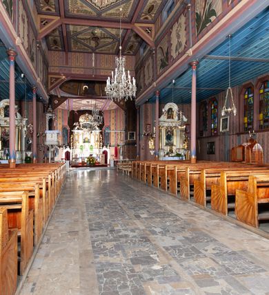 Zdjęcie nr 12: Kościół drewniany, o konstrukcji ramowej, na kamiennej podmurówce, elewacje jednolite, szalowane z listwowaniem o układzie pionowym z gzymsami pseudoarkadkowymi. Wieża zachodnia o konstrukcji słupowej, z szalunkiem o układzie poziomym z fryzem arkadkowym w górnej partii. Więźba dachowa drewniana, krokwiowa. Budynek na betonowym podmurowaniu przesłoniętym daszkiem gontowym. Kościół orientowany, trójnawowy, na rzucie wydłużonego krzyża, z płytkim transeptem, prezbiterium zakończone trójbocznie, z zakrystią przylegającą od południa, na skrzyżowaniu naw kopuła. Od zachodu wieża czterokondygnacyjna, na planie kwadratu przechodzącego w kwadrat o coraz bardziej ściętych narożach, ujęta dwiema kruchtami na rzucie kwadratu. Wnętrze o układzie bazylikowym, nawy podzielone smukłymi, żeliwnymi kolumienkami. Pomiędzy nawą a prezbiterium otwór tęczowy w formie kwadratu o ściętych narożach. Parapet chóru muzycznego w zachodniej części kościoła podparty na czterech kolumienkach. Nawa główna nakryta stropem z widocznymi elementami konstrukcji więźby, nawy boczne przekryte pułapami deskowymi. Posadzka z płyt kamiennych. Elewacja frontowa w formie wieży z dwoma kruchtami. Na każdej z trzech osi wejście z dwuskrzydłowymi drzwiami klepkowymi o układzie rombowym, z oknami w partii szczytu, nakryte daszkami okapowymi. Nad wejściem głównym żeliwna tabliczka z napisem „E(RZ)H(ERZOG) / ALBRECHT / 1888”. Na każdej kondygnacji wieży okna w formie stojącego prostokąta zwieńczone daszkami pulpitowymi, na pierwszej jedno, na drugiej trzy, na trzeciej cztery, na czwartej osiem. W elewacji północnej i południowej cztery podwójne otwory okienne w formie stojącego prostokąta o ściętych górnych narożach w partii naw bocznych oraz pięć okienek w formie leżącego prostokąta w partii nawy głównej. W elewacji południowej wejście z dwuskrzydłowymi drzwiami klepkowymi o układzie rombowym. Na osi skrzydeł transeptu po jednym otworze okiennym w formie stojącego prostokąta o ściętych górnych narożach. Na ścianie wschodniej prezbiterium Ogrójec w formie grupy Ukrzyżowania nakrytej daszkiem pulpitowym, flankowany dwoma otworami okiennymi w kształcie stojącego prostokąta o ściętych górnych narożach. Nawa główna nakryta dachem dwuspadowym. W połowie długości dachu oraz na skrzyżowaniu naw wznoszą się ponad kalenicą wieżyczki na sygnaturkę, na ośmiobocznym obitym blachą trzonie, z latarniami zwieńczonymi cebulastymi hełmami. Nad nawami bocznymi dachy pulpitowe, ramiona transeptu nakrywają dachy naczółkowe. Wieża zwieńczona dachem wieżowym, ośmiobocznym, z iglicą zakończoną krzyżem. 


