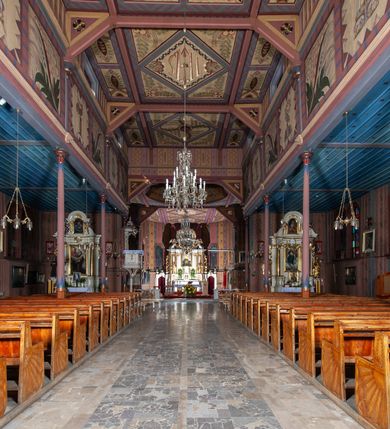 Zdjęcie nr 11: Kościół drewniany, o konstrukcji ramowej, na kamiennej podmurówce, elewacje jednolite, szalowane z listwowaniem o układzie pionowym z gzymsami pseudoarkadkowymi. Wieża zachodnia o konstrukcji słupowej, z szalunkiem o układzie poziomym z fryzem arkadkowym w górnej partii. Więźba dachowa drewniana, krokwiowa. Budynek na betonowym podmurowaniu przesłoniętym daszkiem gontowym. Kościół orientowany, trójnawowy, na rzucie wydłużonego krzyża, z płytkim transeptem, prezbiterium zakończone trójbocznie, z zakrystią przylegającą od południa, na skrzyżowaniu naw kopuła. Od zachodu wieża czterokondygnacyjna, na planie kwadratu przechodzącego w kwadrat o coraz bardziej ściętych narożach, ujęta dwiema kruchtami na rzucie kwadratu. Wnętrze o układzie bazylikowym, nawy podzielone smukłymi, żeliwnymi kolumienkami. Pomiędzy nawą a prezbiterium otwór tęczowy w formie kwadratu o ściętych narożach. Parapet chóru muzycznego w zachodniej części kościoła podparty na czterech kolumienkach. Nawa główna nakryta stropem z widocznymi elementami konstrukcji więźby, nawy boczne przekryte pułapami deskowymi. Posadzka z płyt kamiennych. Elewacja frontowa w formie wieży z dwoma kruchtami. Na każdej z trzech osi wejście z dwuskrzydłowymi drzwiami klepkowymi o układzie rombowym, z oknami w partii szczytu, nakryte daszkami okapowymi. Nad wejściem głównym żeliwna tabliczka z napisem „E(RZ)H(ERZOG) / ALBRECHT / 1888”. Na każdej kondygnacji wieży okna w formie stojącego prostokąta zwieńczone daszkami pulpitowymi, na pierwszej jedno, na drugiej trzy, na trzeciej cztery, na czwartej osiem. W elewacji północnej i południowej cztery podwójne otwory okienne w formie stojącego prostokąta o ściętych górnych narożach w partii naw bocznych oraz pięć okienek w formie leżącego prostokąta w partii nawy głównej. W elewacji południowej wejście z dwuskrzydłowymi drzwiami klepkowymi o układzie rombowym. Na osi skrzydeł transeptu po jednym otworze okiennym w formie stojącego prostokąta o ściętych górnych narożach. Na ścianie wschodniej prezbiterium Ogrójec w formie grupy Ukrzyżowania nakrytej daszkiem pulpitowym, flankowany dwoma otworami okiennymi w kształcie stojącego prostokąta o ściętych górnych narożach. Nawa główna nakryta dachem dwuspadowym. W połowie długości dachu oraz na skrzyżowaniu naw wznoszą się ponad kalenicą wieżyczki na sygnaturkę, na ośmiobocznym obitym blachą trzonie, z latarniami zwieńczonymi cebulastymi hełmami. Nad nawami bocznymi dachy pulpitowe, ramiona transeptu nakrywają dachy naczółkowe. Wieża zwieńczona dachem wieżowym, ośmiobocznym, z iglicą zakończoną krzyżem. 


