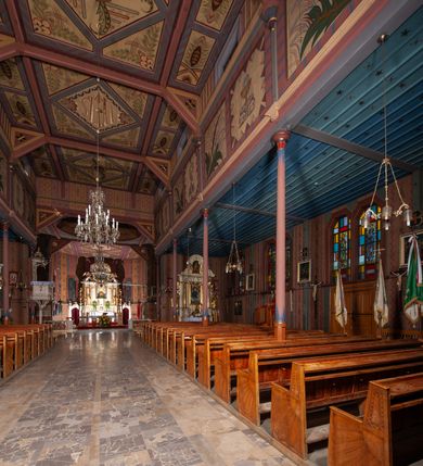 Zdjęcie nr 20: Kościół drewniany, o konstrukcji ramowej, na kamiennej podmurówce, elewacje jednolite, szalowane z listwowaniem o układzie pionowym z gzymsami pseudoarkadkowymi. Wieża zachodnia o konstrukcji słupowej, z szalunkiem o układzie poziomym z fryzem arkadkowym w górnej partii. Więźba dachowa drewniana, krokwiowa. Budynek na betonowym podmurowaniu przesłoniętym daszkiem gontowym. Kościół orientowany, trójnawowy, na rzucie wydłużonego krzyża, z płytkim transeptem, prezbiterium zakończone trójbocznie, z zakrystią przylegającą od południa, na skrzyżowaniu naw kopuła. Od zachodu wieża czterokondygnacyjna, na planie kwadratu przechodzącego w kwadrat o coraz bardziej ściętych narożach, ujęta dwiema kruchtami na rzucie kwadratu. Wnętrze o układzie bazylikowym, nawy podzielone smukłymi, żeliwnymi kolumienkami. Pomiędzy nawą a prezbiterium otwór tęczowy w formie kwadratu o ściętych narożach. Parapet chóru muzycznego w zachodniej części kościoła podparty na czterech kolumienkach. Nawa główna nakryta stropem z widocznymi elementami konstrukcji więźby, nawy boczne przekryte pułapami deskowymi. Posadzka z płyt kamiennych. Elewacja frontowa w formie wieży z dwoma kruchtami. Na każdej z trzech osi wejście z dwuskrzydłowymi drzwiami klepkowymi o układzie rombowym, z oknami w partii szczytu, nakryte daszkami okapowymi. Nad wejściem głównym żeliwna tabliczka z napisem „E(RZ)H(ERZOG) / ALBRECHT / 1888”. Na każdej kondygnacji wieży okna w formie stojącego prostokąta zwieńczone daszkami pulpitowymi, na pierwszej jedno, na drugiej trzy, na trzeciej cztery, na czwartej osiem. W elewacji północnej i południowej cztery podwójne otwory okienne w formie stojącego prostokąta o ściętych górnych narożach w partii naw bocznych oraz pięć okienek w formie leżącego prostokąta w partii nawy głównej. W elewacji południowej wejście z dwuskrzydłowymi drzwiami klepkowymi o układzie rombowym. Na osi skrzydeł transeptu po jednym otworze okiennym w formie stojącego prostokąta o ściętych górnych narożach. Na ścianie wschodniej prezbiterium Ogrójec w formie grupy Ukrzyżowania nakrytej daszkiem pulpitowym, flankowany dwoma otworami okiennymi w kształcie stojącego prostokąta o ściętych górnych narożach. Nawa główna nakryta dachem dwuspadowym. W połowie długości dachu oraz na skrzyżowaniu naw wznoszą się ponad kalenicą wieżyczki na sygnaturkę, na ośmiobocznym obitym blachą trzonie, z latarniami zwieńczonymi cebulastymi hełmami. Nad nawami bocznymi dachy pulpitowe, ramiona transeptu nakrywają dachy naczółkowe. Wieża zwieńczona dachem wieżowym, ośmiobocznym, z iglicą zakończoną krzyżem. 


