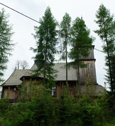 Zdjęcie nr 44: Kościół drewniany o konstrukcji zrębowej, elewacje jednolite, szalowane z listwowaniem, wzmocnione lisicami, wieża o konstrukcji słupowej. Więźba dachowa drewniana, krokwiowa. Budynek na kamiennym podmurowaniu przesłoniętym daszkiem gontowym. Kościół orientowany, jednonawowy z dwiema kaplicami o charakterze transeptu zamkniętymi wielobocznie, na planie krzyża łacińskiego, o węższym prezbiterium zamkniętym trójbocznie. Do wschodniej ściany prezbiterium przylega ogrójec w formie kapliczki, od północy zakrystia na planie prostokąta. Na skrzyżowaniu nawy z południowym ramieniem transeptu kruchta na planie prostokąta. Od zachodu do nawy przylega wieża na planie kwadratu, otoczona sobotami. Prezbiterium i nawa nakryte pozornym sklepieniem tej samej szerokości, obwiedzionym dużym, profilowanym gzymsem podtrzymywanym w nawie przez dwie pary filarów o kanelowanych trzonach, na cokołach. Sklepieniu nawy głównej towarzyszą płaskie i niższe stropy z fasetami w przestrzeniach bocznych. Ściany boczne nawy zwieńczone profilowanym gzymsem. Pomiędzy nawą a prezbiterium otwór tęczowy w formie odcinkowej arkady podtrzymywanej przez filary o kanelowanych trzonach z belką o falistym zarysie. Kaplice boczne otwarte do nawy odcinkowymi arkadami podtrzymywanymi przez filary o kanelowanych trzonach, na cokołach. Parapet chóru muzycznego w zachodniej części nawy, o wybrzuszonej środkowej części, podparty na dwóch filarach o kanelowanych trzonach, na cokołach. Posadzka z płyt ceramicznych. Elewacja frontowa w postaci wieży otoczonej sobotami. Na osi frontowej ściany sobót płytka kruchta zwieńczona gzymsem z przyczółkiem wypełnionym motywem „słoneczka”, przykryta dwuspadowym dachem. Drzwi dwuskrzydłowe, drewniane z okuciami. Korpus wieży podzielony poziomo daszkiem okapowym w dwóch trzecich wysokości, na froncie okno. W dolnej części małe otwory dzwonne z trzech stron, w górnej części duże otwory dzwonne z czterech stron. Wieża zwieńczona hełmem baniastym z latarnią. Ściany sobót szalowane z poziomym listwowaniem, przekryte mansardowym dachem gontowym, na północnej i południowej ścianie dwie pary okien w formie leżącego prostokąta zamkniętego łukiem odcinkowym. W elewacji południowej cztery otwory okienne w formie stojącego prostokąta zamkniętego łukiem odcinkowym, w partii prezbiterium z witrażami. Na elewacji wschodniej ogrójec w formie nadwieszonej kapliczki nakrytej dwuspadowym daszkiem z grupą Ukrzyżowania. W elewacji północnej dwa otwory okienne w formie stojącego prostokąta zamkniętego łukiem odcinkowym. W zakrystii małe kwadratowe okienko. W obu ramionach transeptu analogiczne jak w elewacjach otwory okienne na wschodniej i zachodniej ścianie. Kościół nakryty dachami gontowymi, wyższym nad nawą, niższym nad prezbiterium, dwuspadowym nad nawą oraz wielospadowym nad prezbiterium i ramionami transeptu; nad zakrystią i kruchtą boczną dachy dwuspadowe. W połowie długości dachu, nad nawą, od strony prezbiterium wznosi się ponad kalenicą wieżyczka na sygnaturkę, obita blachą, na ośmiobocznym trzonie, z latarnią zwieńczoną niewielkim baniastym hełmem z latarnią.
