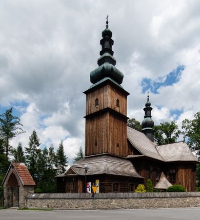 Zdjęcie nr 1: Kościół drewniany o konstrukcji zrębowej, elewacje jednolite, szalowane z listwowaniem, wzmocnione lisicami, wieża o konstrukcji słupowej. Więźba dachowa drewniana, krokwiowa. Budynek na kamiennym podmurowaniu przesłoniętym daszkiem gontowym. Kościół orientowany, jednonawowy z dwiema kaplicami o charakterze transeptu zamkniętymi wielobocznie, na planie krzyża łacińskiego, o węższym prezbiterium zamkniętym trójbocznie. Do wschodniej ściany prezbiterium przylega ogrójec w formie kapliczki, od północy zakrystia na planie prostokąta. Na skrzyżowaniu nawy z południowym ramieniem transeptu kruchta na planie prostokąta. Od zachodu do nawy przylega wieża na planie kwadratu, otoczona sobotami. Prezbiterium i nawa nakryte pozornym sklepieniem tej samej szerokości, obwiedzionym dużym, profilowanym gzymsem podtrzymywanym w nawie przez dwie pary filarów o kanelowanych trzonach, na cokołach. Sklepieniu nawy głównej towarzyszą płaskie i niższe stropy z fasetami w przestrzeniach bocznych. Ściany boczne nawy zwieńczone profilowanym gzymsem. Pomiędzy nawą a prezbiterium otwór tęczowy w formie odcinkowej arkady podtrzymywanej przez filary o kanelowanych trzonach z belką o falistym zarysie. Kaplice boczne otwarte do nawy odcinkowymi arkadami podtrzymywanymi przez filary o kanelowanych trzonach, na cokołach. Parapet chóru muzycznego w zachodniej części nawy, o wybrzuszonej środkowej części, podparty na dwóch filarach o kanelowanych trzonach, na cokołach. Posadzka z płyt ceramicznych. Elewacja frontowa w postaci wieży otoczonej sobotami. Na osi frontowej ściany sobót płytka kruchta zwieńczona gzymsem z przyczółkiem wypełnionym motywem „słoneczka”, przykryta dwuspadowym dachem. Drzwi dwuskrzydłowe, drewniane z okuciami. Korpus wieży podzielony poziomo daszkiem okapowym w dwóch trzecich wysokości, na froncie okno. W dolnej części małe otwory dzwonne z trzech stron, w górnej części duże otwory dzwonne z czterech stron. Wieża zwieńczona hełmem baniastym z latarnią. Ściany sobót szalowane z poziomym listwowaniem, przekryte mansardowym dachem gontowym, na północnej i południowej ścianie dwie pary okien w formie leżącego prostokąta zamkniętego łukiem odcinkowym. W elewacji południowej cztery otwory okienne w formie stojącego prostokąta zamkniętego łukiem odcinkowym, w partii prezbiterium z witrażami. Na elewacji wschodniej ogrójec w formie nadwieszonej kapliczki nakrytej dwuspadowym daszkiem z grupą Ukrzyżowania. W elewacji północnej dwa otwory okienne w formie stojącego prostokąta zamkniętego łukiem odcinkowym. W zakrystii małe kwadratowe okienko. W obu ramionach transeptu analogiczne jak w elewacjach otwory okienne na wschodniej i zachodniej ścianie. Kościół nakryty dachami gontowymi, wyższym nad nawą, niższym nad prezbiterium, dwuspadowym nad nawą oraz wielospadowym nad prezbiterium i ramionami transeptu; nad zakrystią i kruchtą boczną dachy dwuspadowe. W połowie długości dachu, nad nawą, od strony prezbiterium wznosi się ponad kalenicą wieżyczka na sygnaturkę, obita blachą, na ośmiobocznym trzonie, z latarnią zwieńczoną niewielkim baniastym hełmem z latarnią.
