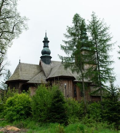 Zdjęcie nr 43: Kościół drewniany o konstrukcji zrębowej, elewacje jednolite, szalowane z listwowaniem, wzmocnione lisicami, wieża o konstrukcji słupowej. Więźba dachowa drewniana, krokwiowa. Budynek na kamiennym podmurowaniu przesłoniętym daszkiem gontowym. Kościół orientowany, jednonawowy z dwiema kaplicami o charakterze transeptu zamkniętymi wielobocznie, na planie krzyża łacińskiego, o węższym prezbiterium zamkniętym trójbocznie. Do wschodniej ściany prezbiterium przylega ogrójec w formie kapliczki, od północy zakrystia na planie prostokąta. Na skrzyżowaniu nawy z południowym ramieniem transeptu kruchta na planie prostokąta. Od zachodu do nawy przylega wieża na planie kwadratu, otoczona sobotami. Prezbiterium i nawa nakryte pozornym sklepieniem tej samej szerokości, obwiedzionym dużym, profilowanym gzymsem podtrzymywanym w nawie przez dwie pary filarów o kanelowanych trzonach, na cokołach. Sklepieniu nawy głównej towarzyszą płaskie i niższe stropy z fasetami w przestrzeniach bocznych. Ściany boczne nawy zwieńczone profilowanym gzymsem. Pomiędzy nawą a prezbiterium otwór tęczowy w formie odcinkowej arkady podtrzymywanej przez filary o kanelowanych trzonach z belką o falistym zarysie. Kaplice boczne otwarte do nawy odcinkowymi arkadami podtrzymywanymi przez filary o kanelowanych trzonach, na cokołach. Parapet chóru muzycznego w zachodniej części nawy, o wybrzuszonej środkowej części, podparty na dwóch filarach o kanelowanych trzonach, na cokołach. Posadzka z płyt ceramicznych. Elewacja frontowa w postaci wieży otoczonej sobotami. Na osi frontowej ściany sobót płytka kruchta zwieńczona gzymsem z przyczółkiem wypełnionym motywem „słoneczka”, przykryta dwuspadowym dachem. Drzwi dwuskrzydłowe, drewniane z okuciami. Korpus wieży podzielony poziomo daszkiem okapowym w dwóch trzecich wysokości, na froncie okno. W dolnej części małe otwory dzwonne z trzech stron, w górnej części duże otwory dzwonne z czterech stron. Wieża zwieńczona hełmem baniastym z latarnią. Ściany sobót szalowane z poziomym listwowaniem, przekryte mansardowym dachem gontowym, na północnej i południowej ścianie dwie pary okien w formie leżącego prostokąta zamkniętego łukiem odcinkowym. W elewacji południowej cztery otwory okienne w formie stojącego prostokąta zamkniętego łukiem odcinkowym, w partii prezbiterium z witrażami. Na elewacji wschodniej ogrójec w formie nadwieszonej kapliczki nakrytej dwuspadowym daszkiem z grupą Ukrzyżowania. W elewacji północnej dwa otwory okienne w formie stojącego prostokąta zamkniętego łukiem odcinkowym. W zakrystii małe kwadratowe okienko. W obu ramionach transeptu analogiczne jak w elewacjach otwory okienne na wschodniej i zachodniej ścianie. Kościół nakryty dachami gontowymi, wyższym nad nawą, niższym nad prezbiterium, dwuspadowym nad nawą oraz wielospadowym nad prezbiterium i ramionami transeptu; nad zakrystią i kruchtą boczną dachy dwuspadowe. W połowie długości dachu, nad nawą, od strony prezbiterium wznosi się ponad kalenicą wieżyczka na sygnaturkę, obita blachą, na ośmiobocznym trzonie, z latarnią zwieńczoną niewielkim baniastym hełmem z latarnią.
