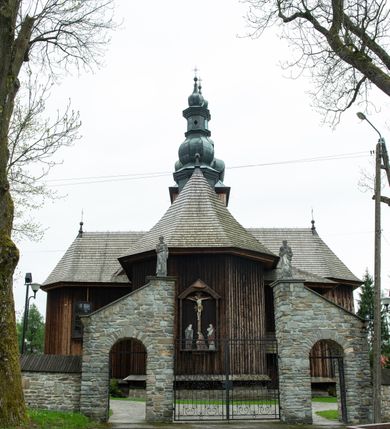 Zdjęcie nr 53: Kościół drewniany o konstrukcji zrębowej, elewacje jednolite, szalowane z listwowaniem, wzmocnione lisicami, wieża o konstrukcji słupowej. Więźba dachowa drewniana, krokwiowa. Budynek na kamiennym podmurowaniu przesłoniętym daszkiem gontowym. Kościół orientowany, jednonawowy z dwiema kaplicami o charakterze transeptu zamkniętymi wielobocznie, na planie krzyża łacińskiego, o węższym prezbiterium zamkniętym trójbocznie. Do wschodniej ściany prezbiterium przylega ogrójec w formie kapliczki, od północy zakrystia na planie prostokąta. Na skrzyżowaniu nawy z południowym ramieniem transeptu kruchta na planie prostokąta. Od zachodu do nawy przylega wieża na planie kwadratu, otoczona sobotami. Prezbiterium i nawa nakryte pozornym sklepieniem tej samej szerokości, obwiedzionym dużym, profilowanym gzymsem podtrzymywanym w nawie przez dwie pary filarów o kanelowanych trzonach, na cokołach. Sklepieniu nawy głównej towarzyszą płaskie i niższe stropy z fasetami w przestrzeniach bocznych. Ściany boczne nawy zwieńczone profilowanym gzymsem. Pomiędzy nawą a prezbiterium otwór tęczowy w formie odcinkowej arkady podtrzymywanej przez filary o kanelowanych trzonach z belką o falistym zarysie. Kaplice boczne otwarte do nawy odcinkowymi arkadami podtrzymywanymi przez filary o kanelowanych trzonach, na cokołach. Parapet chóru muzycznego w zachodniej części nawy, o wybrzuszonej środkowej części, podparty na dwóch filarach o kanelowanych trzonach, na cokołach. Posadzka z płyt ceramicznych. Elewacja frontowa w postaci wieży otoczonej sobotami. Na osi frontowej ściany sobót płytka kruchta zwieńczona gzymsem z przyczółkiem wypełnionym motywem „słoneczka”, przykryta dwuspadowym dachem. Drzwi dwuskrzydłowe, drewniane z okuciami. Korpus wieży podzielony poziomo daszkiem okapowym w dwóch trzecich wysokości, na froncie okno. W dolnej części małe otwory dzwonne z trzech stron, w górnej części duże otwory dzwonne z czterech stron. Wieża zwieńczona hełmem baniastym z latarnią. Ściany sobót szalowane z poziomym listwowaniem, przekryte mansardowym dachem gontowym, na północnej i południowej ścianie dwie pary okien w formie leżącego prostokąta zamkniętego łukiem odcinkowym. W elewacji południowej cztery otwory okienne w formie stojącego prostokąta zamkniętego łukiem odcinkowym, w partii prezbiterium z witrażami. Na elewacji wschodniej ogrójec w formie nadwieszonej kapliczki nakrytej dwuspadowym daszkiem z grupą Ukrzyżowania. W elewacji północnej dwa otwory okienne w formie stojącego prostokąta zamkniętego łukiem odcinkowym. W zakrystii małe kwadratowe okienko. W obu ramionach transeptu analogiczne jak w elewacjach otwory okienne na wschodniej i zachodniej ścianie. Kościół nakryty dachami gontowymi, wyższym nad nawą, niższym nad prezbiterium, dwuspadowym nad nawą oraz wielospadowym nad prezbiterium i ramionami transeptu; nad zakrystią i kruchtą boczną dachy dwuspadowe. W połowie długości dachu, nad nawą, od strony prezbiterium wznosi się ponad kalenicą wieżyczka na sygnaturkę, obita blachą, na ośmiobocznym trzonie, z latarnią zwieńczoną niewielkim baniastym hełmem z latarnią.
