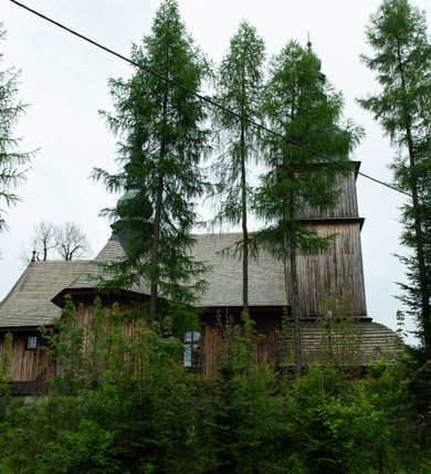 Zdjęcie nr 45: Kościół drewniany o konstrukcji zrębowej, elewacje jednolite, szalowane z listwowaniem, wzmocnione lisicami, wieża o konstrukcji słupowej. Więźba dachowa drewniana, krokwiowa. Budynek na kamiennym podmurowaniu przesłoniętym daszkiem gontowym. Kościół orientowany, jednonawowy z dwiema kaplicami o charakterze transeptu zamkniętymi wielobocznie, na planie krzyża łacińskiego, o węższym prezbiterium zamkniętym trójbocznie. Do wschodniej ściany prezbiterium przylega ogrójec w formie kapliczki, od północy zakrystia na planie prostokąta. Na skrzyżowaniu nawy z południowym ramieniem transeptu kruchta na planie prostokąta. Od zachodu do nawy przylega wieża na planie kwadratu, otoczona sobotami. Prezbiterium i nawa nakryte pozornym sklepieniem tej samej szerokości, obwiedzionym dużym, profilowanym gzymsem podtrzymywanym w nawie przez dwie pary filarów o kanelowanych trzonach, na cokołach. Sklepieniu nawy głównej towarzyszą płaskie i niższe stropy z fasetami w przestrzeniach bocznych. Ściany boczne nawy zwieńczone profilowanym gzymsem. Pomiędzy nawą a prezbiterium otwór tęczowy w formie odcinkowej arkady podtrzymywanej przez filary o kanelowanych trzonach z belką o falistym zarysie. Kaplice boczne otwarte do nawy odcinkowymi arkadami podtrzymywanymi przez filary o kanelowanych trzonach, na cokołach. Parapet chóru muzycznego w zachodniej części nawy, o wybrzuszonej środkowej części, podparty na dwóch filarach o kanelowanych trzonach, na cokołach. Posadzka z płyt ceramicznych. Elewacja frontowa w postaci wieży otoczonej sobotami. Na osi frontowej ściany sobót płytka kruchta zwieńczona gzymsem z przyczółkiem wypełnionym motywem „słoneczka”, przykryta dwuspadowym dachem. Drzwi dwuskrzydłowe, drewniane z okuciami. Korpus wieży podzielony poziomo daszkiem okapowym w dwóch trzecich wysokości, na froncie okno. W dolnej części małe otwory dzwonne z trzech stron, w górnej części duże otwory dzwonne z czterech stron. Wieża zwieńczona hełmem baniastym z latarnią. Ściany sobót szalowane z poziomym listwowaniem, przekryte mansardowym dachem gontowym, na północnej i południowej ścianie dwie pary okien w formie leżącego prostokąta zamkniętego łukiem odcinkowym. W elewacji południowej cztery otwory okienne w formie stojącego prostokąta zamkniętego łukiem odcinkowym, w partii prezbiterium z witrażami. Na elewacji wschodniej ogrójec w formie nadwieszonej kapliczki nakrytej dwuspadowym daszkiem z grupą Ukrzyżowania. W elewacji północnej dwa otwory okienne w formie stojącego prostokąta zamkniętego łukiem odcinkowym. W zakrystii małe kwadratowe okienko. W obu ramionach transeptu analogiczne jak w elewacjach otwory okienne na wschodniej i zachodniej ścianie. Kościół nakryty dachami gontowymi, wyższym nad nawą, niższym nad prezbiterium, dwuspadowym nad nawą oraz wielospadowym nad prezbiterium i ramionami transeptu; nad zakrystią i kruchtą boczną dachy dwuspadowe. W połowie długości dachu, nad nawą, od strony prezbiterium wznosi się ponad kalenicą wieżyczka na sygnaturkę, obita blachą, na ośmiobocznym trzonie, z latarnią zwieńczoną niewielkim baniastym hełmem z latarnią.
