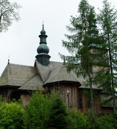 Zdjęcie nr 42: Kościół drewniany o konstrukcji zrębowej, elewacje jednolite, szalowane z listwowaniem, wzmocnione lisicami, wieża o konstrukcji słupowej. Więźba dachowa drewniana, krokwiowa. Budynek na kamiennym podmurowaniu przesłoniętym daszkiem gontowym. Kościół orientowany, jednonawowy z dwiema kaplicami o charakterze transeptu zamkniętymi wielobocznie, na planie krzyża łacińskiego, o węższym prezbiterium zamkniętym trójbocznie. Do wschodniej ściany prezbiterium przylega ogrójec w formie kapliczki, od północy zakrystia na planie prostokąta. Na skrzyżowaniu nawy z południowym ramieniem transeptu kruchta na planie prostokąta. Od zachodu do nawy przylega wieża na planie kwadratu, otoczona sobotami. Prezbiterium i nawa nakryte pozornym sklepieniem tej samej szerokości, obwiedzionym dużym, profilowanym gzymsem podtrzymywanym w nawie przez dwie pary filarów o kanelowanych trzonach, na cokołach. Sklepieniu nawy głównej towarzyszą płaskie i niższe stropy z fasetami w przestrzeniach bocznych. Ściany boczne nawy zwieńczone profilowanym gzymsem. Pomiędzy nawą a prezbiterium otwór tęczowy w formie odcinkowej arkady podtrzymywanej przez filary o kanelowanych trzonach z belką o falistym zarysie. Kaplice boczne otwarte do nawy odcinkowymi arkadami podtrzymywanymi przez filary o kanelowanych trzonach, na cokołach. Parapet chóru muzycznego w zachodniej części nawy, o wybrzuszonej środkowej części, podparty na dwóch filarach o kanelowanych trzonach, na cokołach. Posadzka z płyt ceramicznych. Elewacja frontowa w postaci wieży otoczonej sobotami. Na osi frontowej ściany sobót płytka kruchta zwieńczona gzymsem z przyczółkiem wypełnionym motywem „słoneczka”, przykryta dwuspadowym dachem. Drzwi dwuskrzydłowe, drewniane z okuciami. Korpus wieży podzielony poziomo daszkiem okapowym w dwóch trzecich wysokości, na froncie okno. W dolnej części małe otwory dzwonne z trzech stron, w górnej części duże otwory dzwonne z czterech stron. Wieża zwieńczona hełmem baniastym z latarnią. Ściany sobót szalowane z poziomym listwowaniem, przekryte mansardowym dachem gontowym, na północnej i południowej ścianie dwie pary okien w formie leżącego prostokąta zamkniętego łukiem odcinkowym. W elewacji południowej cztery otwory okienne w formie stojącego prostokąta zamkniętego łukiem odcinkowym, w partii prezbiterium z witrażami. Na elewacji wschodniej ogrójec w formie nadwieszonej kapliczki nakrytej dwuspadowym daszkiem z grupą Ukrzyżowania. W elewacji północnej dwa otwory okienne w formie stojącego prostokąta zamkniętego łukiem odcinkowym. W zakrystii małe kwadratowe okienko. W obu ramionach transeptu analogiczne jak w elewacjach otwory okienne na wschodniej i zachodniej ścianie. Kościół nakryty dachami gontowymi, wyższym nad nawą, niższym nad prezbiterium, dwuspadowym nad nawą oraz wielospadowym nad prezbiterium i ramionami transeptu; nad zakrystią i kruchtą boczną dachy dwuspadowe. W połowie długości dachu, nad nawą, od strony prezbiterium wznosi się ponad kalenicą wieżyczka na sygnaturkę, obita blachą, na ośmiobocznym trzonie, z latarnią zwieńczoną niewielkim baniastym hełmem z latarnią.
