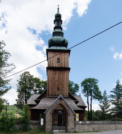 Zdjęcie nr 17: Kościół drewniany o konstrukcji zrębowej, elewacje jednolite, szalowane z listwowaniem, wzmocnione lisicami, wieża o konstrukcji słupowej. Więźba dachowa drewniana, krokwiowa. Budynek na kamiennym podmurowaniu przesłoniętym daszkiem gontowym. Kościół orientowany, jednonawowy z dwiema kaplicami o charakterze transeptu zamkniętymi wielobocznie, na planie krzyża łacińskiego, o węższym prezbiterium zamkniętym trójbocznie. Do wschodniej ściany prezbiterium przylega ogrójec w formie kapliczki, od północy zakrystia na planie prostokąta. Na skrzyżowaniu nawy z południowym ramieniem transeptu kruchta na planie prostokąta. Od zachodu do nawy przylega wieża na planie kwadratu, otoczona sobotami. Prezbiterium i nawa nakryte pozornym sklepieniem tej samej szerokości, obwiedzionym dużym, profilowanym gzymsem podtrzymywanym w nawie przez dwie pary filarów o kanelowanych trzonach, na cokołach. Sklepieniu nawy głównej towarzyszą płaskie i niższe stropy z fasetami w przestrzeniach bocznych. Ściany boczne nawy zwieńczone profilowanym gzymsem. Pomiędzy nawą a prezbiterium otwór tęczowy w formie odcinkowej arkady podtrzymywanej przez filary o kanelowanych trzonach z belką o falistym zarysie. Kaplice boczne otwarte do nawy odcinkowymi arkadami podtrzymywanymi przez filary o kanelowanych trzonach, na cokołach. Parapet chóru muzycznego w zachodniej części nawy, o wybrzuszonej środkowej części, podparty na dwóch filarach o kanelowanych trzonach, na cokołach. Posadzka z płyt ceramicznych. Elewacja frontowa w postaci wieży otoczonej sobotami. Na osi frontowej ściany sobót płytka kruchta zwieńczona gzymsem z przyczółkiem wypełnionym motywem „słoneczka”, przykryta dwuspadowym dachem. Drzwi dwuskrzydłowe, drewniane z okuciami. Korpus wieży podzielony poziomo daszkiem okapowym w dwóch trzecich wysokości, na froncie okno. W dolnej części małe otwory dzwonne z trzech stron, w górnej części duże otwory dzwonne z czterech stron. Wieża zwieńczona hełmem baniastym z latarnią. Ściany sobót szalowane z poziomym listwowaniem, przekryte mansardowym dachem gontowym, na północnej i południowej ścianie dwie pary okien w formie leżącego prostokąta zamkniętego łukiem odcinkowym. W elewacji południowej cztery otwory okienne w formie stojącego prostokąta zamkniętego łukiem odcinkowym, w partii prezbiterium z witrażami. Na elewacji wschodniej ogrójec w formie nadwieszonej kapliczki nakrytej dwuspadowym daszkiem z grupą Ukrzyżowania. W elewacji północnej dwa otwory okienne w formie stojącego prostokąta zamkniętego łukiem odcinkowym. W zakrystii małe kwadratowe okienko. W obu ramionach transeptu analogiczne jak w elewacjach otwory okienne na wschodniej i zachodniej ścianie. Kościół nakryty dachami gontowymi, wyższym nad nawą, niższym nad prezbiterium, dwuspadowym nad nawą oraz wielospadowym nad prezbiterium i ramionami transeptu; nad zakrystią i kruchtą boczną dachy dwuspadowe. W połowie długości dachu, nad nawą, od strony prezbiterium wznosi się ponad kalenicą wieżyczka na sygnaturkę, obita blachą, na ośmiobocznym trzonie, z latarnią zwieńczoną niewielkim baniastym hełmem z latarnią.
