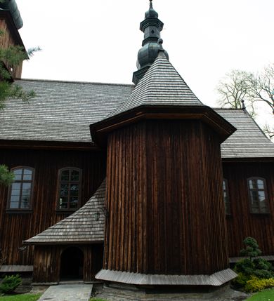 Zdjęcie nr 37: Kościół drewniany o konstrukcji zrębowej, elewacje jednolite, szalowane z listwowaniem, wzmocnione lisicami, wieża o konstrukcji słupowej. Więźba dachowa drewniana, krokwiowa. Budynek na kamiennym podmurowaniu przesłoniętym daszkiem gontowym. Kościół orientowany, jednonawowy z dwiema kaplicami o charakterze transeptu zamkniętymi wielobocznie, na planie krzyża łacińskiego, o węższym prezbiterium zamkniętym trójbocznie. Do wschodniej ściany prezbiterium przylega ogrójec w formie kapliczki, od północy zakrystia na planie prostokąta. Na skrzyżowaniu nawy z południowym ramieniem transeptu kruchta na planie prostokąta. Od zachodu do nawy przylega wieża na planie kwadratu, otoczona sobotami. Prezbiterium i nawa nakryte pozornym sklepieniem tej samej szerokości, obwiedzionym dużym, profilowanym gzymsem podtrzymywanym w nawie przez dwie pary filarów o kanelowanych trzonach, na cokołach. Sklepieniu nawy głównej towarzyszą płaskie i niższe stropy z fasetami w przestrzeniach bocznych. Ściany boczne nawy zwieńczone profilowanym gzymsem. Pomiędzy nawą a prezbiterium otwór tęczowy w formie odcinkowej arkady podtrzymywanej przez filary o kanelowanych trzonach z belką o falistym zarysie. Kaplice boczne otwarte do nawy odcinkowymi arkadami podtrzymywanymi przez filary o kanelowanych trzonach, na cokołach. Parapet chóru muzycznego w zachodniej części nawy, o wybrzuszonej środkowej części, podparty na dwóch filarach o kanelowanych trzonach, na cokołach. Posadzka z płyt ceramicznych. Elewacja frontowa w postaci wieży otoczonej sobotami. Na osi frontowej ściany sobót płytka kruchta zwieńczona gzymsem z przyczółkiem wypełnionym motywem „słoneczka”, przykryta dwuspadowym dachem. Drzwi dwuskrzydłowe, drewniane z okuciami. Korpus wieży podzielony poziomo daszkiem okapowym w dwóch trzecich wysokości, na froncie okno. W dolnej części małe otwory dzwonne z trzech stron, w górnej części duże otwory dzwonne z czterech stron. Wieża zwieńczona hełmem baniastym z latarnią. Ściany sobót szalowane z poziomym listwowaniem, przekryte mansardowym dachem gontowym, na północnej i południowej ścianie dwie pary okien w formie leżącego prostokąta zamkniętego łukiem odcinkowym. W elewacji południowej cztery otwory okienne w formie stojącego prostokąta zamkniętego łukiem odcinkowym, w partii prezbiterium z witrażami. Na elewacji wschodniej ogrójec w formie nadwieszonej kapliczki nakrytej dwuspadowym daszkiem z grupą Ukrzyżowania. W elewacji północnej dwa otwory okienne w formie stojącego prostokąta zamkniętego łukiem odcinkowym. W zakrystii małe kwadratowe okienko. W obu ramionach transeptu analogiczne jak w elewacjach otwory okienne na wschodniej i zachodniej ścianie. Kościół nakryty dachami gontowymi, wyższym nad nawą, niższym nad prezbiterium, dwuspadowym nad nawą oraz wielospadowym nad prezbiterium i ramionami transeptu; nad zakrystią i kruchtą boczną dachy dwuspadowe. W połowie długości dachu, nad nawą, od strony prezbiterium wznosi się ponad kalenicą wieżyczka na sygnaturkę, obita blachą, na ośmiobocznym trzonie, z latarnią zwieńczoną niewielkim baniastym hełmem z latarnią.
