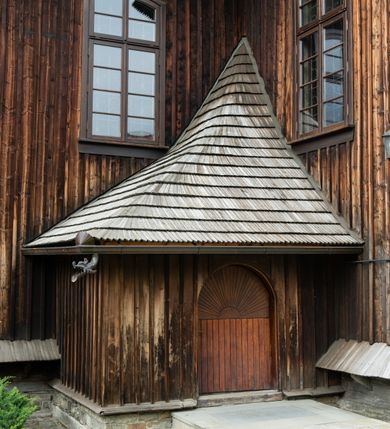 Zdjęcie nr 38: Kościół drewniany o konstrukcji zrębowej, elewacje jednolite, szalowane z listwowaniem, wzmocnione lisicami, wieża o konstrukcji słupowej. Więźba dachowa drewniana, krokwiowa. Budynek na kamiennym podmurowaniu przesłoniętym daszkiem gontowym. Kościół orientowany, jednonawowy z dwiema kaplicami o charakterze transeptu zamkniętymi wielobocznie, na planie krzyża łacińskiego, o węższym prezbiterium zamkniętym trójbocznie. Do wschodniej ściany prezbiterium przylega ogrójec w formie kapliczki, od północy zakrystia na planie prostokąta. Na skrzyżowaniu nawy z południowym ramieniem transeptu kruchta na planie prostokąta. Od zachodu do nawy przylega wieża na planie kwadratu, otoczona sobotami. Prezbiterium i nawa nakryte pozornym sklepieniem tej samej szerokości, obwiedzionym dużym, profilowanym gzymsem podtrzymywanym w nawie przez dwie pary filarów o kanelowanych trzonach, na cokołach. Sklepieniu nawy głównej towarzyszą płaskie i niższe stropy z fasetami w przestrzeniach bocznych. Ściany boczne nawy zwieńczone profilowanym gzymsem. Pomiędzy nawą a prezbiterium otwór tęczowy w formie odcinkowej arkady podtrzymywanej przez filary o kanelowanych trzonach z belką o falistym zarysie. Kaplice boczne otwarte do nawy odcinkowymi arkadami podtrzymywanymi przez filary o kanelowanych trzonach, na cokołach. Parapet chóru muzycznego w zachodniej części nawy, o wybrzuszonej środkowej części, podparty na dwóch filarach o kanelowanych trzonach, na cokołach. Posadzka z płyt ceramicznych. Elewacja frontowa w postaci wieży otoczonej sobotami. Na osi frontowej ściany sobót płytka kruchta zwieńczona gzymsem z przyczółkiem wypełnionym motywem „słoneczka”, przykryta dwuspadowym dachem. Drzwi dwuskrzydłowe, drewniane z okuciami. Korpus wieży podzielony poziomo daszkiem okapowym w dwóch trzecich wysokości, na froncie okno. W dolnej części małe otwory dzwonne z trzech stron, w górnej części duże otwory dzwonne z czterech stron. Wieża zwieńczona hełmem baniastym z latarnią. Ściany sobót szalowane z poziomym listwowaniem, przekryte mansardowym dachem gontowym, na północnej i południowej ścianie dwie pary okien w formie leżącego prostokąta zamkniętego łukiem odcinkowym. W elewacji południowej cztery otwory okienne w formie stojącego prostokąta zamkniętego łukiem odcinkowym, w partii prezbiterium z witrażami. Na elewacji wschodniej ogrójec w formie nadwieszonej kapliczki nakrytej dwuspadowym daszkiem z grupą Ukrzyżowania. W elewacji północnej dwa otwory okienne w formie stojącego prostokąta zamkniętego łukiem odcinkowym. W zakrystii małe kwadratowe okienko. W obu ramionach transeptu analogiczne jak w elewacjach otwory okienne na wschodniej i zachodniej ścianie. Kościół nakryty dachami gontowymi, wyższym nad nawą, niższym nad prezbiterium, dwuspadowym nad nawą oraz wielospadowym nad prezbiterium i ramionami transeptu; nad zakrystią i kruchtą boczną dachy dwuspadowe. W połowie długości dachu, nad nawą, od strony prezbiterium wznosi się ponad kalenicą wieżyczka na sygnaturkę, obita blachą, na ośmiobocznym trzonie, z latarnią zwieńczoną niewielkim baniastym hełmem z latarnią.
