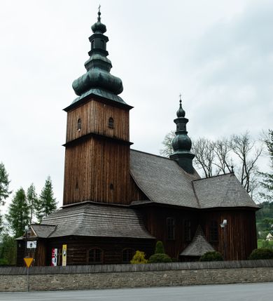 Zdjęcie nr 22: Kościół drewniany o konstrukcji zrębowej, elewacje jednolite, szalowane z listwowaniem, wzmocnione lisicami, wieża o konstrukcji słupowej. Więźba dachowa drewniana, krokwiowa. Budynek na kamiennym podmurowaniu przesłoniętym daszkiem gontowym. Kościół orientowany, jednonawowy z dwiema kaplicami o charakterze transeptu zamkniętymi wielobocznie, na planie krzyża łacińskiego, o węższym prezbiterium zamkniętym trójbocznie. Do wschodniej ściany prezbiterium przylega ogrójec w formie kapliczki, od północy zakrystia na planie prostokąta. Na skrzyżowaniu nawy z południowym ramieniem transeptu kruchta na planie prostokąta. Od zachodu do nawy przylega wieża na planie kwadratu, otoczona sobotami. Prezbiterium i nawa nakryte pozornym sklepieniem tej samej szerokości, obwiedzionym dużym, profilowanym gzymsem podtrzymywanym w nawie przez dwie pary filarów o kanelowanych trzonach, na cokołach. Sklepieniu nawy głównej towarzyszą płaskie i niższe stropy z fasetami w przestrzeniach bocznych. Ściany boczne nawy zwieńczone profilowanym gzymsem. Pomiędzy nawą a prezbiterium otwór tęczowy w formie odcinkowej arkady podtrzymywanej przez filary o kanelowanych trzonach z belką o falistym zarysie. Kaplice boczne otwarte do nawy odcinkowymi arkadami podtrzymywanymi przez filary o kanelowanych trzonach, na cokołach. Parapet chóru muzycznego w zachodniej części nawy, o wybrzuszonej środkowej części, podparty na dwóch filarach o kanelowanych trzonach, na cokołach. Posadzka z płyt ceramicznych. Elewacja frontowa w postaci wieży otoczonej sobotami. Na osi frontowej ściany sobót płytka kruchta zwieńczona gzymsem z przyczółkiem wypełnionym motywem „słoneczka”, przykryta dwuspadowym dachem. Drzwi dwuskrzydłowe, drewniane z okuciami. Korpus wieży podzielony poziomo daszkiem okapowym w dwóch trzecich wysokości, na froncie okno. W dolnej części małe otwory dzwonne z trzech stron, w górnej części duże otwory dzwonne z czterech stron. Wieża zwieńczona hełmem baniastym z latarnią. Ściany sobót szalowane z poziomym listwowaniem, przekryte mansardowym dachem gontowym, na północnej i południowej ścianie dwie pary okien w formie leżącego prostokąta zamkniętego łukiem odcinkowym. W elewacji południowej cztery otwory okienne w formie stojącego prostokąta zamkniętego łukiem odcinkowym, w partii prezbiterium z witrażami. Na elewacji wschodniej ogrójec w formie nadwieszonej kapliczki nakrytej dwuspadowym daszkiem z grupą Ukrzyżowania. W elewacji północnej dwa otwory okienne w formie stojącego prostokąta zamkniętego łukiem odcinkowym. W zakrystii małe kwadratowe okienko. W obu ramionach transeptu analogiczne jak w elewacjach otwory okienne na wschodniej i zachodniej ścianie. Kościół nakryty dachami gontowymi, wyższym nad nawą, niższym nad prezbiterium, dwuspadowym nad nawą oraz wielospadowym nad prezbiterium i ramionami transeptu; nad zakrystią i kruchtą boczną dachy dwuspadowe. W połowie długości dachu, nad nawą, od strony prezbiterium wznosi się ponad kalenicą wieżyczka na sygnaturkę, obita blachą, na ośmiobocznym trzonie, z latarnią zwieńczoną niewielkim baniastym hełmem z latarnią.

