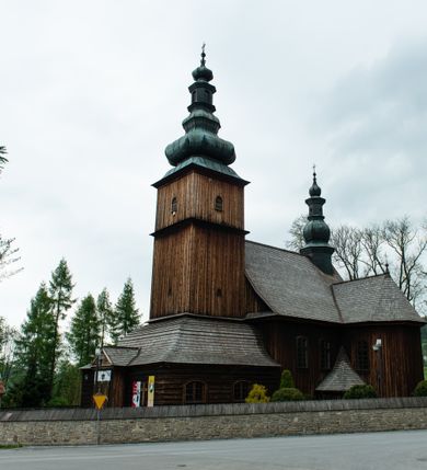 Zdjęcie nr 21: Kościół drewniany o konstrukcji zrębowej, elewacje jednolite, szalowane z listwowaniem, wzmocnione lisicami, wieża o konstrukcji słupowej. Więźba dachowa drewniana, krokwiowa. Budynek na kamiennym podmurowaniu przesłoniętym daszkiem gontowym. Kościół orientowany, jednonawowy z dwiema kaplicami o charakterze transeptu zamkniętymi wielobocznie, na planie krzyża łacińskiego, o węższym prezbiterium zamkniętym trójbocznie. Do wschodniej ściany prezbiterium przylega ogrójec w formie kapliczki, od północy zakrystia na planie prostokąta. Na skrzyżowaniu nawy z południowym ramieniem transeptu kruchta na planie prostokąta. Od zachodu do nawy przylega wieża na planie kwadratu, otoczona sobotami. Prezbiterium i nawa nakryte pozornym sklepieniem tej samej szerokości, obwiedzionym dużym, profilowanym gzymsem podtrzymywanym w nawie przez dwie pary filarów o kanelowanych trzonach, na cokołach. Sklepieniu nawy głównej towarzyszą płaskie i niższe stropy z fasetami w przestrzeniach bocznych. Ściany boczne nawy zwieńczone profilowanym gzymsem. Pomiędzy nawą a prezbiterium otwór tęczowy w formie odcinkowej arkady podtrzymywanej przez filary o kanelowanych trzonach z belką o falistym zarysie. Kaplice boczne otwarte do nawy odcinkowymi arkadami podtrzymywanymi przez filary o kanelowanych trzonach, na cokołach. Parapet chóru muzycznego w zachodniej części nawy, o wybrzuszonej środkowej części, podparty na dwóch filarach o kanelowanych trzonach, na cokołach. Posadzka z płyt ceramicznych. Elewacja frontowa w postaci wieży otoczonej sobotami. Na osi frontowej ściany sobót płytka kruchta zwieńczona gzymsem z przyczółkiem wypełnionym motywem „słoneczka”, przykryta dwuspadowym dachem. Drzwi dwuskrzydłowe, drewniane z okuciami. Korpus wieży podzielony poziomo daszkiem okapowym w dwóch trzecich wysokości, na froncie okno. W dolnej części małe otwory dzwonne z trzech stron, w górnej części duże otwory dzwonne z czterech stron. Wieża zwieńczona hełmem baniastym z latarnią. Ściany sobót szalowane z poziomym listwowaniem, przekryte mansardowym dachem gontowym, na północnej i południowej ścianie dwie pary okien w formie leżącego prostokąta zamkniętego łukiem odcinkowym. W elewacji południowej cztery otwory okienne w formie stojącego prostokąta zamkniętego łukiem odcinkowym, w partii prezbiterium z witrażami. Na elewacji wschodniej ogrójec w formie nadwieszonej kapliczki nakrytej dwuspadowym daszkiem z grupą Ukrzyżowania. W elewacji północnej dwa otwory okienne w formie stojącego prostokąta zamkniętego łukiem odcinkowym. W zakrystii małe kwadratowe okienko. W obu ramionach transeptu analogiczne jak w elewacjach otwory okienne na wschodniej i zachodniej ścianie. Kościół nakryty dachami gontowymi, wyższym nad nawą, niższym nad prezbiterium, dwuspadowym nad nawą oraz wielospadowym nad prezbiterium i ramionami transeptu; nad zakrystią i kruchtą boczną dachy dwuspadowe. W połowie długości dachu, nad nawą, od strony prezbiterium wznosi się ponad kalenicą wieżyczka na sygnaturkę, obita blachą, na ośmiobocznym trzonie, z latarnią zwieńczoną niewielkim baniastym hełmem z latarnią.
