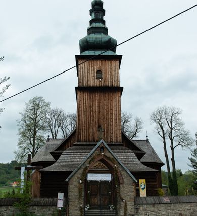 Zdjęcie nr 18: Kościół drewniany o konstrukcji zrębowej, elewacje jednolite, szalowane z listwowaniem, wzmocnione lisicami, wieża o konstrukcji słupowej. Więźba dachowa drewniana, krokwiowa. Budynek na kamiennym podmurowaniu przesłoniętym daszkiem gontowym. Kościół orientowany, jednonawowy z dwiema kaplicami o charakterze transeptu zamkniętymi wielobocznie, na planie krzyża łacińskiego, o węższym prezbiterium zamkniętym trójbocznie. Do wschodniej ściany prezbiterium przylega ogrójec w formie kapliczki, od północy zakrystia na planie prostokąta. Na skrzyżowaniu nawy z południowym ramieniem transeptu kruchta na planie prostokąta. Od zachodu do nawy przylega wieża na planie kwadratu, otoczona sobotami. Prezbiterium i nawa nakryte pozornym sklepieniem tej samej szerokości, obwiedzionym dużym, profilowanym gzymsem podtrzymywanym w nawie przez dwie pary filarów o kanelowanych trzonach, na cokołach. Sklepieniu nawy głównej towarzyszą płaskie i niższe stropy z fasetami w przestrzeniach bocznych. Ściany boczne nawy zwieńczone profilowanym gzymsem. Pomiędzy nawą a prezbiterium otwór tęczowy w formie odcinkowej arkady podtrzymywanej przez filary o kanelowanych trzonach z belką o falistym zarysie. Kaplice boczne otwarte do nawy odcinkowymi arkadami podtrzymywanymi przez filary o kanelowanych trzonach, na cokołach. Parapet chóru muzycznego w zachodniej części nawy, o wybrzuszonej środkowej części, podparty na dwóch filarach o kanelowanych trzonach, na cokołach. Posadzka z płyt ceramicznych. Elewacja frontowa w postaci wieży otoczonej sobotami. Na osi frontowej ściany sobót płytka kruchta zwieńczona gzymsem z przyczółkiem wypełnionym motywem „słoneczka”, przykryta dwuspadowym dachem. Drzwi dwuskrzydłowe, drewniane z okuciami. Korpus wieży podzielony poziomo daszkiem okapowym w dwóch trzecich wysokości, na froncie okno. W dolnej części małe otwory dzwonne z trzech stron, w górnej części duże otwory dzwonne z czterech stron. Wieża zwieńczona hełmem baniastym z latarnią. Ściany sobót szalowane z poziomym listwowaniem, przekryte mansardowym dachem gontowym, na północnej i południowej ścianie dwie pary okien w formie leżącego prostokąta zamkniętego łukiem odcinkowym. W elewacji południowej cztery otwory okienne w formie stojącego prostokąta zamkniętego łukiem odcinkowym, w partii prezbiterium z witrażami. Na elewacji wschodniej ogrójec w formie nadwieszonej kapliczki nakrytej dwuspadowym daszkiem z grupą Ukrzyżowania. W elewacji północnej dwa otwory okienne w formie stojącego prostokąta zamkniętego łukiem odcinkowym. W zakrystii małe kwadratowe okienko. W obu ramionach transeptu analogiczne jak w elewacjach otwory okienne na wschodniej i zachodniej ścianie. Kościół nakryty dachami gontowymi, wyższym nad nawą, niższym nad prezbiterium, dwuspadowym nad nawą oraz wielospadowym nad prezbiterium i ramionami transeptu; nad zakrystią i kruchtą boczną dachy dwuspadowe. W połowie długości dachu, nad nawą, od strony prezbiterium wznosi się ponad kalenicą wieżyczka na sygnaturkę, obita blachą, na ośmiobocznym trzonie, z latarnią zwieńczoną niewielkim baniastym hełmem z latarnią.
