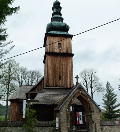 Zdjęcie nr 19: Kościół drewniany o konstrukcji zrębowej, elewacje jednolite, szalowane z listwowaniem, wzmocnione lisicami, wieża o konstrukcji słupowej. Więźba dachowa drewniana, krokwiowa. Budynek na kamiennym podmurowaniu przesłoniętym daszkiem gontowym. Kościół orientowany, jednonawowy z dwiema kaplicami o charakterze transeptu zamkniętymi wielobocznie, na planie krzyża łacińskiego, o węższym prezbiterium zamkniętym trójbocznie. Do wschodniej ściany prezbiterium przylega ogrójec w formie kapliczki, od północy zakrystia na planie prostokąta. Na skrzyżowaniu nawy z południowym ramieniem transeptu kruchta na planie prostokąta. Od zachodu do nawy przylega wieża na planie kwadratu, otoczona sobotami. Prezbiterium i nawa nakryte pozornym sklepieniem tej samej szerokości, obwiedzionym dużym, profilowanym gzymsem podtrzymywanym w nawie przez dwie pary filarów o kanelowanych trzonach, na cokołach. Sklepieniu nawy głównej towarzyszą płaskie i niższe stropy z fasetami w przestrzeniach bocznych. Ściany boczne nawy zwieńczone profilowanym gzymsem. Pomiędzy nawą a prezbiterium otwór tęczowy w formie odcinkowej arkady podtrzymywanej przez filary o kanelowanych trzonach z belką o falistym zarysie. Kaplice boczne otwarte do nawy odcinkowymi arkadami podtrzymywanymi przez filary o kanelowanych trzonach, na cokołach. Parapet chóru muzycznego w zachodniej części nawy, o wybrzuszonej środkowej części, podparty na dwóch filarach o kanelowanych trzonach, na cokołach. Posadzka z płyt ceramicznych. Elewacja frontowa w postaci wieży otoczonej sobotami. Na osi frontowej ściany sobót płytka kruchta zwieńczona gzymsem z przyczółkiem wypełnionym motywem „słoneczka”, przykryta dwuspadowym dachem. Drzwi dwuskrzydłowe, drewniane z okuciami. Korpus wieży podzielony poziomo daszkiem okapowym w dwóch trzecich wysokości, na froncie okno. W dolnej części małe otwory dzwonne z trzech stron, w górnej części duże otwory dzwonne z czterech stron. Wieża zwieńczona hełmem baniastym z latarnią. Ściany sobót szalowane z poziomym listwowaniem, przekryte mansardowym dachem gontowym, na północnej i południowej ścianie dwie pary okien w formie leżącego prostokąta zamkniętego łukiem odcinkowym. W elewacji południowej cztery otwory okienne w formie stojącego prostokąta zamkniętego łukiem odcinkowym, w partii prezbiterium z witrażami. Na elewacji wschodniej ogrójec w formie nadwieszonej kapliczki nakrytej dwuspadowym daszkiem z grupą Ukrzyżowania. W elewacji północnej dwa otwory okienne w formie stojącego prostokąta zamkniętego łukiem odcinkowym. W zakrystii małe kwadratowe okienko. W obu ramionach transeptu analogiczne jak w elewacjach otwory okienne na wschodniej i zachodniej ścianie. Kościół nakryty dachami gontowymi, wyższym nad nawą, niższym nad prezbiterium, dwuspadowym nad nawą oraz wielospadowym nad prezbiterium i ramionami transeptu; nad zakrystią i kruchtą boczną dachy dwuspadowe. W połowie długości dachu, nad nawą, od strony prezbiterium wznosi się ponad kalenicą wieżyczka na sygnaturkę, obita blachą, na ośmiobocznym trzonie, z latarnią zwieńczoną niewielkim baniastym hełmem z latarnią.
