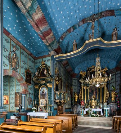 Zdjęcie nr 71: Kościół drewniany o konstrukcji zrębowej, elewacje jednolite, szalowane z listwowaniem, wzmocnione lisicami, wieża o konstrukcji słupowej. Więźba dachowa drewniana, krokwiowa. Budynek na kamiennym podmurowaniu przesłoniętym daszkiem gontowym. Kościół orientowany, jednonawowy z dwiema kaplicami o charakterze transeptu zamkniętymi wielobocznie, na planie krzyża łacińskiego, o węższym prezbiterium zamkniętym trójbocznie. Do wschodniej ściany prezbiterium przylega ogrójec w formie kapliczki, od północy zakrystia na planie prostokąta. Na skrzyżowaniu nawy z południowym ramieniem transeptu kruchta na planie prostokąta. Od zachodu do nawy przylega wieża na planie kwadratu, otoczona sobotami. Prezbiterium i nawa nakryte pozornym sklepieniem tej samej szerokości, obwiedzionym dużym, profilowanym gzymsem podtrzymywanym w nawie przez dwie pary filarów o kanelowanych trzonach, na cokołach. Sklepieniu nawy głównej towarzyszą płaskie i niższe stropy z fasetami w przestrzeniach bocznych. Ściany boczne nawy zwieńczone profilowanym gzymsem. Pomiędzy nawą a prezbiterium otwór tęczowy w formie odcinkowej arkady podtrzymywanej przez filary o kanelowanych trzonach z belką o falistym zarysie. Kaplice boczne otwarte do nawy odcinkowymi arkadami podtrzymywanymi przez filary o kanelowanych trzonach, na cokołach. Parapet chóru muzycznego w zachodniej części nawy, o wybrzuszonej środkowej części, podparty na dwóch filarach o kanelowanych trzonach, na cokołach. Posadzka z płyt ceramicznych. Elewacja frontowa w postaci wieży otoczonej sobotami. Na osi frontowej ściany sobót płytka kruchta zwieńczona gzymsem z przyczółkiem wypełnionym motywem „słoneczka”, przykryta dwuspadowym dachem. Drzwi dwuskrzydłowe, drewniane z okuciami. Korpus wieży podzielony poziomo daszkiem okapowym w dwóch trzecich wysokości, na froncie okno. W dolnej części małe otwory dzwonne z trzech stron, w górnej części duże otwory dzwonne z czterech stron. Wieża zwieńczona hełmem baniastym z latarnią. Ściany sobót szalowane z poziomym listwowaniem, przekryte mansardowym dachem gontowym, na północnej i południowej ścianie dwie pary okien w formie leżącego prostokąta zamkniętego łukiem odcinkowym. W elewacji południowej cztery otwory okienne w formie stojącego prostokąta zamkniętego łukiem odcinkowym, w partii prezbiterium z witrażami. Na elewacji wschodniej ogrójec w formie nadwieszonej kapliczki nakrytej dwuspadowym daszkiem z grupą Ukrzyżowania. W elewacji północnej dwa otwory okienne w formie stojącego prostokąta zamkniętego łukiem odcinkowym. W zakrystii małe kwadratowe okienko. W obu ramionach transeptu analogiczne jak w elewacjach otwory okienne na wschodniej i zachodniej ścianie. Kościół nakryty dachami gontowymi, wyższym nad nawą, niższym nad prezbiterium, dwuspadowym nad nawą oraz wielospadowym nad prezbiterium i ramionami transeptu; nad zakrystią i kruchtą boczną dachy dwuspadowe. W połowie długości dachu, nad nawą, od strony prezbiterium wznosi się ponad kalenicą wieżyczka na sygnaturkę, obita blachą, na ośmiobocznym trzonie, z latarnią zwieńczoną niewielkim baniastym hełmem z latarnią.
