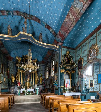 Zdjęcie nr 72: Kościół drewniany o konstrukcji zrębowej, elewacje jednolite, szalowane z listwowaniem, wzmocnione lisicami, wieża o konstrukcji słupowej. Więźba dachowa drewniana, krokwiowa. Budynek na kamiennym podmurowaniu przesłoniętym daszkiem gontowym. Kościół orientowany, jednonawowy z dwiema kaplicami o charakterze transeptu zamkniętymi wielobocznie, na planie krzyża łacińskiego, o węższym prezbiterium zamkniętym trójbocznie. Do wschodniej ściany prezbiterium przylega ogrójec w formie kapliczki, od północy zakrystia na planie prostokąta. Na skrzyżowaniu nawy z południowym ramieniem transeptu kruchta na planie prostokąta. Od zachodu do nawy przylega wieża na planie kwadratu, otoczona sobotami. Prezbiterium i nawa nakryte pozornym sklepieniem tej samej szerokości, obwiedzionym dużym, profilowanym gzymsem podtrzymywanym w nawie przez dwie pary filarów o kanelowanych trzonach, na cokołach. Sklepieniu nawy głównej towarzyszą płaskie i niższe stropy z fasetami w przestrzeniach bocznych. Ściany boczne nawy zwieńczone profilowanym gzymsem. Pomiędzy nawą a prezbiterium otwór tęczowy w formie odcinkowej arkady podtrzymywanej przez filary o kanelowanych trzonach z belką o falistym zarysie. Kaplice boczne otwarte do nawy odcinkowymi arkadami podtrzymywanymi przez filary o kanelowanych trzonach, na cokołach. Parapet chóru muzycznego w zachodniej części nawy, o wybrzuszonej środkowej części, podparty na dwóch filarach o kanelowanych trzonach, na cokołach. Posadzka z płyt ceramicznych. Elewacja frontowa w postaci wieży otoczonej sobotami. Na osi frontowej ściany sobót płytka kruchta zwieńczona gzymsem z przyczółkiem wypełnionym motywem „słoneczka”, przykryta dwuspadowym dachem. Drzwi dwuskrzydłowe, drewniane z okuciami. Korpus wieży podzielony poziomo daszkiem okapowym w dwóch trzecich wysokości, na froncie okno. W dolnej części małe otwory dzwonne z trzech stron, w górnej części duże otwory dzwonne z czterech stron. Wieża zwieńczona hełmem baniastym z latarnią. Ściany sobót szalowane z poziomym listwowaniem, przekryte mansardowym dachem gontowym, na północnej i południowej ścianie dwie pary okien w formie leżącego prostokąta zamkniętego łukiem odcinkowym. W elewacji południowej cztery otwory okienne w formie stojącego prostokąta zamkniętego łukiem odcinkowym, w partii prezbiterium z witrażami. Na elewacji wschodniej ogrójec w formie nadwieszonej kapliczki nakrytej dwuspadowym daszkiem z grupą Ukrzyżowania. W elewacji północnej dwa otwory okienne w formie stojącego prostokąta zamkniętego łukiem odcinkowym. W zakrystii małe kwadratowe okienko. W obu ramionach transeptu analogiczne jak w elewacjach otwory okienne na wschodniej i zachodniej ścianie. Kościół nakryty dachami gontowymi, wyższym nad nawą, niższym nad prezbiterium, dwuspadowym nad nawą oraz wielospadowym nad prezbiterium i ramionami transeptu; nad zakrystią i kruchtą boczną dachy dwuspadowe. W połowie długości dachu, nad nawą, od strony prezbiterium wznosi się ponad kalenicą wieżyczka na sygnaturkę, obita blachą, na ośmiobocznym trzonie, z latarnią zwieńczoną niewielkim baniastym hełmem z latarnią.
