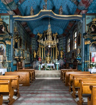 Zdjęcie nr 68: Kościół drewniany o konstrukcji zrębowej, elewacje jednolite, szalowane z listwowaniem, wzmocnione lisicami, wieża o konstrukcji słupowej. Więźba dachowa drewniana, krokwiowa. Budynek na kamiennym podmurowaniu przesłoniętym daszkiem gontowym. Kościół orientowany, jednonawowy z dwiema kaplicami o charakterze transeptu zamkniętymi wielobocznie, na planie krzyża łacińskiego, o węższym prezbiterium zamkniętym trójbocznie. Do wschodniej ściany prezbiterium przylega ogrójec w formie kapliczki, od północy zakrystia na planie prostokąta. Na skrzyżowaniu nawy z południowym ramieniem transeptu kruchta na planie prostokąta. Od zachodu do nawy przylega wieża na planie kwadratu, otoczona sobotami. Prezbiterium i nawa nakryte pozornym sklepieniem tej samej szerokości, obwiedzionym dużym, profilowanym gzymsem podtrzymywanym w nawie przez dwie pary filarów o kanelowanych trzonach, na cokołach. Sklepieniu nawy głównej towarzyszą płaskie i niższe stropy z fasetami w przestrzeniach bocznych. Ściany boczne nawy zwieńczone profilowanym gzymsem. Pomiędzy nawą a prezbiterium otwór tęczowy w formie odcinkowej arkady podtrzymywanej przez filary o kanelowanych trzonach z belką o falistym zarysie. Kaplice boczne otwarte do nawy odcinkowymi arkadami podtrzymywanymi przez filary o kanelowanych trzonach, na cokołach. Parapet chóru muzycznego w zachodniej części nawy, o wybrzuszonej środkowej części, podparty na dwóch filarach o kanelowanych trzonach, na cokołach. Posadzka z płyt ceramicznych. Elewacja frontowa w postaci wieży otoczonej sobotami. Na osi frontowej ściany sobót płytka kruchta zwieńczona gzymsem z przyczółkiem wypełnionym motywem „słoneczka”, przykryta dwuspadowym dachem. Drzwi dwuskrzydłowe, drewniane z okuciami. Korpus wieży podzielony poziomo daszkiem okapowym w dwóch trzecich wysokości, na froncie okno. W dolnej części małe otwory dzwonne z trzech stron, w górnej części duże otwory dzwonne z czterech stron. Wieża zwieńczona hełmem baniastym z latarnią. Ściany sobót szalowane z poziomym listwowaniem, przekryte mansardowym dachem gontowym, na północnej i południowej ścianie dwie pary okien w formie leżącego prostokąta zamkniętego łukiem odcinkowym. W elewacji południowej cztery otwory okienne w formie stojącego prostokąta zamkniętego łukiem odcinkowym, w partii prezbiterium z witrażami. Na elewacji wschodniej ogrójec w formie nadwieszonej kapliczki nakrytej dwuspadowym daszkiem z grupą Ukrzyżowania. W elewacji północnej dwa otwory okienne w formie stojącego prostokąta zamkniętego łukiem odcinkowym. W zakrystii małe kwadratowe okienko. W obu ramionach transeptu analogiczne jak w elewacjach otwory okienne na wschodniej i zachodniej ścianie. Kościół nakryty dachami gontowymi, wyższym nad nawą, niższym nad prezbiterium, dwuspadowym nad nawą oraz wielospadowym nad prezbiterium i ramionami transeptu; nad zakrystią i kruchtą boczną dachy dwuspadowe. W połowie długości dachu, nad nawą, od strony prezbiterium wznosi się ponad kalenicą wieżyczka na sygnaturkę, obita blachą, na ośmiobocznym trzonie, z latarnią zwieńczoną niewielkim baniastym hełmem z latarnią.
