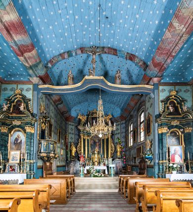 Zdjęcie nr 67: Kościół drewniany o konstrukcji zrębowej, elewacje jednolite, szalowane z listwowaniem, wzmocnione lisicami, wieża o konstrukcji słupowej. Więźba dachowa drewniana, krokwiowa. Budynek na kamiennym podmurowaniu przesłoniętym daszkiem gontowym. Kościół orientowany, jednonawowy z dwiema kaplicami o charakterze transeptu zamkniętymi wielobocznie, na planie krzyża łacińskiego, o węższym prezbiterium zamkniętym trójbocznie. Do wschodniej ściany prezbiterium przylega ogrójec w formie kapliczki, od północy zakrystia na planie prostokąta. Na skrzyżowaniu nawy z południowym ramieniem transeptu kruchta na planie prostokąta. Od zachodu do nawy przylega wieża na planie kwadratu, otoczona sobotami. Prezbiterium i nawa nakryte pozornym sklepieniem tej samej szerokości, obwiedzionym dużym, profilowanym gzymsem podtrzymywanym w nawie przez dwie pary filarów o kanelowanych trzonach, na cokołach. Sklepieniu nawy głównej towarzyszą płaskie i niższe stropy z fasetami w przestrzeniach bocznych. Ściany boczne nawy zwieńczone profilowanym gzymsem. Pomiędzy nawą a prezbiterium otwór tęczowy w formie odcinkowej arkady podtrzymywanej przez filary o kanelowanych trzonach z belką o falistym zarysie. Kaplice boczne otwarte do nawy odcinkowymi arkadami podtrzymywanymi przez filary o kanelowanych trzonach, na cokołach. Parapet chóru muzycznego w zachodniej części nawy, o wybrzuszonej środkowej części, podparty na dwóch filarach o kanelowanych trzonach, na cokołach. Posadzka z płyt ceramicznych. Elewacja frontowa w postaci wieży otoczonej sobotami. Na osi frontowej ściany sobót płytka kruchta zwieńczona gzymsem z przyczółkiem wypełnionym motywem „słoneczka”, przykryta dwuspadowym dachem. Drzwi dwuskrzydłowe, drewniane z okuciami. Korpus wieży podzielony poziomo daszkiem okapowym w dwóch trzecich wysokości, na froncie okno. W dolnej części małe otwory dzwonne z trzech stron, w górnej części duże otwory dzwonne z czterech stron. Wieża zwieńczona hełmem baniastym z latarnią. Ściany sobót szalowane z poziomym listwowaniem, przekryte mansardowym dachem gontowym, na północnej i południowej ścianie dwie pary okien w formie leżącego prostokąta zamkniętego łukiem odcinkowym. W elewacji południowej cztery otwory okienne w formie stojącego prostokąta zamkniętego łukiem odcinkowym, w partii prezbiterium z witrażami. Na elewacji wschodniej ogrójec w formie nadwieszonej kapliczki nakrytej dwuspadowym daszkiem z grupą Ukrzyżowania. W elewacji północnej dwa otwory okienne w formie stojącego prostokąta zamkniętego łukiem odcinkowym. W zakrystii małe kwadratowe okienko. W obu ramionach transeptu analogiczne jak w elewacjach otwory okienne na wschodniej i zachodniej ścianie. Kościół nakryty dachami gontowymi, wyższym nad nawą, niższym nad prezbiterium, dwuspadowym nad nawą oraz wielospadowym nad prezbiterium i ramionami transeptu; nad zakrystią i kruchtą boczną dachy dwuspadowe. W połowie długości dachu, nad nawą, od strony prezbiterium wznosi się ponad kalenicą wieżyczka na sygnaturkę, obita blachą, na ośmiobocznym trzonie, z latarnią zwieńczoną niewielkim baniastym hełmem z latarnią.
