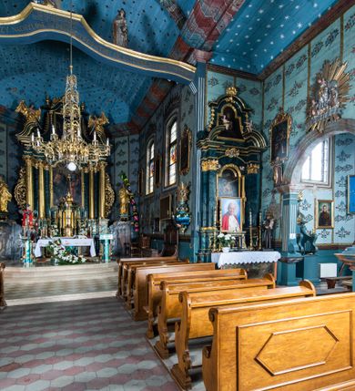 Zdjęcie nr 2: Kościół drewniany o konstrukcji zrębowej, elewacje jednolite, szalowane z listwowaniem, wzmocnione lisicami, wieża o konstrukcji słupowej. Więźba dachowa drewniana, krokwiowa. Budynek na kamiennym podmurowaniu przesłoniętym daszkiem gontowym. Kościół orientowany, jednonawowy z dwiema kaplicami o charakterze transeptu zamkniętymi wielobocznie, na planie krzyża łacińskiego, o węższym prezbiterium zamkniętym trójbocznie. Do wschodniej ściany prezbiterium przylega ogrójec w formie kapliczki, od północy zakrystia na planie prostokąta. Na skrzyżowaniu nawy z południowym ramieniem transeptu kruchta na planie prostokąta. Od zachodu do nawy przylega wieża na planie kwadratu, otoczona sobotami. Prezbiterium i nawa nakryte pozornym sklepieniem tej samej szerokości, obwiedzionym dużym, profilowanym gzymsem podtrzymywanym w nawie przez dwie pary filarów o kanelowanych trzonach, na cokołach. Sklepieniu nawy głównej towarzyszą płaskie i niższe stropy z fasetami w przestrzeniach bocznych. Ściany boczne nawy zwieńczone profilowanym gzymsem. Pomiędzy nawą a prezbiterium otwór tęczowy w formie odcinkowej arkady podtrzymywanej przez filary o kanelowanych trzonach z belką o falistym zarysie. Kaplice boczne otwarte do nawy odcinkowymi arkadami podtrzymywanymi przez filary o kanelowanych trzonach, na cokołach. Parapet chóru muzycznego w zachodniej części nawy, o wybrzuszonej środkowej części, podparty na dwóch filarach o kanelowanych trzonach, na cokołach. Posadzka z płyt ceramicznych. Elewacja frontowa w postaci wieży otoczonej sobotami. Na osi frontowej ściany sobót płytka kruchta zwieńczona gzymsem z przyczółkiem wypełnionym motywem „słoneczka”, przykryta dwuspadowym dachem. Drzwi dwuskrzydłowe, drewniane z okuciami. Korpus wieży podzielony poziomo daszkiem okapowym w dwóch trzecich wysokości, na froncie okno. W dolnej części małe otwory dzwonne z trzech stron, w górnej części duże otwory dzwonne z czterech stron. Wieża zwieńczona hełmem baniastym z latarnią. Ściany sobót szalowane z poziomym listwowaniem, przekryte mansardowym dachem gontowym, na północnej i południowej ścianie dwie pary okien w formie leżącego prostokąta zamkniętego łukiem odcinkowym. W elewacji południowej cztery otwory okienne w formie stojącego prostokąta zamkniętego łukiem odcinkowym, w partii prezbiterium z witrażami. Na elewacji wschodniej ogrójec w formie nadwieszonej kapliczki nakrytej dwuspadowym daszkiem z grupą Ukrzyżowania. W elewacji północnej dwa otwory okienne w formie stojącego prostokąta zamkniętego łukiem odcinkowym. W zakrystii małe kwadratowe okienko. W obu ramionach transeptu analogiczne jak w elewacjach otwory okienne na wschodniej i zachodniej ścianie. Kościół nakryty dachami gontowymi, wyższym nad nawą, niższym nad prezbiterium, dwuspadowym nad nawą oraz wielospadowym nad prezbiterium i ramionami transeptu; nad zakrystią i kruchtą boczną dachy dwuspadowe. W połowie długości dachu, nad nawą, od strony prezbiterium wznosi się ponad kalenicą wieżyczka na sygnaturkę, obita blachą, na ośmiobocznym trzonie, z latarnią zwieńczoną niewielkim baniastym hełmem z latarnią.
