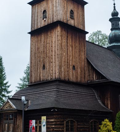 Zdjęcie nr 31: Kościół drewniany o konstrukcji zrębowej, elewacje jednolite, szalowane z listwowaniem, wzmocnione lisicami, wieża o konstrukcji słupowej. Więźba dachowa drewniana, krokwiowa. Budynek na kamiennym podmurowaniu przesłoniętym daszkiem gontowym. Kościół orientowany, jednonawowy z dwiema kaplicami o charakterze transeptu zamkniętymi wielobocznie, na planie krzyża łacińskiego, o węższym prezbiterium zamkniętym trójbocznie. Do wschodniej ściany prezbiterium przylega ogrójec w formie kapliczki, od północy zakrystia na planie prostokąta. Na skrzyżowaniu nawy z południowym ramieniem transeptu kruchta na planie prostokąta. Od zachodu do nawy przylega wieża na planie kwadratu, otoczona sobotami. Prezbiterium i nawa nakryte pozornym sklepieniem tej samej szerokości, obwiedzionym dużym, profilowanym gzymsem podtrzymywanym w nawie przez dwie pary filarów o kanelowanych trzonach, na cokołach. Sklepieniu nawy głównej towarzyszą płaskie i niższe stropy z fasetami w przestrzeniach bocznych. Ściany boczne nawy zwieńczone profilowanym gzymsem. Pomiędzy nawą a prezbiterium otwór tęczowy w formie odcinkowej arkady podtrzymywanej przez filary o kanelowanych trzonach z belką o falistym zarysie. Kaplice boczne otwarte do nawy odcinkowymi arkadami podtrzymywanymi przez filary o kanelowanych trzonach, na cokołach. Parapet chóru muzycznego w zachodniej części nawy, o wybrzuszonej środkowej części, podparty na dwóch filarach o kanelowanych trzonach, na cokołach. Posadzka z płyt ceramicznych. Elewacja frontowa w postaci wieży otoczonej sobotami. Na osi frontowej ściany sobót płytka kruchta zwieńczona gzymsem z przyczółkiem wypełnionym motywem „słoneczka”, przykryta dwuspadowym dachem. Drzwi dwuskrzydłowe, drewniane z okuciami. Korpus wieży podzielony poziomo daszkiem okapowym w dwóch trzecich wysokości, na froncie okno. W dolnej części małe otwory dzwonne z trzech stron, w górnej części duże otwory dzwonne z czterech stron. Wieża zwieńczona hełmem baniastym z latarnią. Ściany sobót szalowane z poziomym listwowaniem, przekryte mansardowym dachem gontowym, na północnej i południowej ścianie dwie pary okien w formie leżącego prostokąta zamkniętego łukiem odcinkowym. W elewacji południowej cztery otwory okienne w formie stojącego prostokąta zamkniętego łukiem odcinkowym, w partii prezbiterium z witrażami. Na elewacji wschodniej ogrójec w formie nadwieszonej kapliczki nakrytej dwuspadowym daszkiem z grupą Ukrzyżowania. W elewacji północnej dwa otwory okienne w formie stojącego prostokąta zamkniętego łukiem odcinkowym. W zakrystii małe kwadratowe okienko. W obu ramionach transeptu analogiczne jak w elewacjach otwory okienne na wschodniej i zachodniej ścianie. Kościół nakryty dachami gontowymi, wyższym nad nawą, niższym nad prezbiterium, dwuspadowym nad nawą oraz wielospadowym nad prezbiterium i ramionami transeptu; nad zakrystią i kruchtą boczną dachy dwuspadowe. W połowie długości dachu, nad nawą, od strony prezbiterium wznosi się ponad kalenicą wieżyczka na sygnaturkę, obita blachą, na ośmiobocznym trzonie, z latarnią zwieńczoną niewielkim baniastym hełmem z latarnią.
