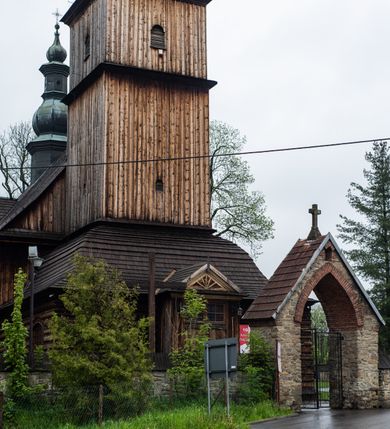 Zdjęcie nr 29: Kościół drewniany o konstrukcji zrębowej, elewacje jednolite, szalowane z listwowaniem, wzmocnione lisicami, wieża o konstrukcji słupowej. Więźba dachowa drewniana, krokwiowa. Budynek na kamiennym podmurowaniu przesłoniętym daszkiem gontowym. Kościół orientowany, jednonawowy z dwiema kaplicami o charakterze transeptu zamkniętymi wielobocznie, na planie krzyża łacińskiego, o węższym prezbiterium zamkniętym trójbocznie. Do wschodniej ściany prezbiterium przylega ogrójec w formie kapliczki, od północy zakrystia na planie prostokąta. Na skrzyżowaniu nawy z południowym ramieniem transeptu kruchta na planie prostokąta. Od zachodu do nawy przylega wieża na planie kwadratu, otoczona sobotami. Prezbiterium i nawa nakryte pozornym sklepieniem tej samej szerokości, obwiedzionym dużym, profilowanym gzymsem podtrzymywanym w nawie przez dwie pary filarów o kanelowanych trzonach, na cokołach. Sklepieniu nawy głównej towarzyszą płaskie i niższe stropy z fasetami w przestrzeniach bocznych. Ściany boczne nawy zwieńczone profilowanym gzymsem. Pomiędzy nawą a prezbiterium otwór tęczowy w formie odcinkowej arkady podtrzymywanej przez filary o kanelowanych trzonach z belką o falistym zarysie. Kaplice boczne otwarte do nawy odcinkowymi arkadami podtrzymywanymi przez filary o kanelowanych trzonach, na cokołach. Parapet chóru muzycznego w zachodniej części nawy, o wybrzuszonej środkowej części, podparty na dwóch filarach o kanelowanych trzonach, na cokołach. Posadzka z płyt ceramicznych. Elewacja frontowa w postaci wieży otoczonej sobotami. Na osi frontowej ściany sobót płytka kruchta zwieńczona gzymsem z przyczółkiem wypełnionym motywem „słoneczka”, przykryta dwuspadowym dachem. Drzwi dwuskrzydłowe, drewniane z okuciami. Korpus wieży podzielony poziomo daszkiem okapowym w dwóch trzecich wysokości, na froncie okno. W dolnej części małe otwory dzwonne z trzech stron, w górnej części duże otwory dzwonne z czterech stron. Wieża zwieńczona hełmem baniastym z latarnią. Ściany sobót szalowane z poziomym listwowaniem, przekryte mansardowym dachem gontowym, na północnej i południowej ścianie dwie pary okien w formie leżącego prostokąta zamkniętego łukiem odcinkowym. W elewacji południowej cztery otwory okienne w formie stojącego prostokąta zamkniętego łukiem odcinkowym, w partii prezbiterium z witrażami. Na elewacji wschodniej ogrójec w formie nadwieszonej kapliczki nakrytej dwuspadowym daszkiem z grupą Ukrzyżowania. W elewacji północnej dwa otwory okienne w formie stojącego prostokąta zamkniętego łukiem odcinkowym. W zakrystii małe kwadratowe okienko. W obu ramionach transeptu analogiczne jak w elewacjach otwory okienne na wschodniej i zachodniej ścianie. Kościół nakryty dachami gontowymi, wyższym nad nawą, niższym nad prezbiterium, dwuspadowym nad nawą oraz wielospadowym nad prezbiterium i ramionami transeptu; nad zakrystią i kruchtą boczną dachy dwuspadowe. W połowie długości dachu, nad nawą, od strony prezbiterium wznosi się ponad kalenicą wieżyczka na sygnaturkę, obita blachą, na ośmiobocznym trzonie, z latarnią zwieńczoną niewielkim baniastym hełmem z latarnią.
