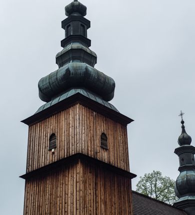 Zdjęcie nr 33: Kościół drewniany o konstrukcji zrębowej, elewacje jednolite, szalowane z listwowaniem, wzmocnione lisicami, wieża o konstrukcji słupowej. Więźba dachowa drewniana, krokwiowa. Budynek na kamiennym podmurowaniu przesłoniętym daszkiem gontowym. Kościół orientowany, jednonawowy z dwiema kaplicami o charakterze transeptu zamkniętymi wielobocznie, na planie krzyża łacińskiego, o węższym prezbiterium zamkniętym trójbocznie. Do wschodniej ściany prezbiterium przylega ogrójec w formie kapliczki, od północy zakrystia na planie prostokąta. Na skrzyżowaniu nawy z południowym ramieniem transeptu kruchta na planie prostokąta. Od zachodu do nawy przylega wieża na planie kwadratu, otoczona sobotami. Prezbiterium i nawa nakryte pozornym sklepieniem tej samej szerokości, obwiedzionym dużym, profilowanym gzymsem podtrzymywanym w nawie przez dwie pary filarów o kanelowanych trzonach, na cokołach. Sklepieniu nawy głównej towarzyszą płaskie i niższe stropy z fasetami w przestrzeniach bocznych. Ściany boczne nawy zwieńczone profilowanym gzymsem. Pomiędzy nawą a prezbiterium otwór tęczowy w formie odcinkowej arkady podtrzymywanej przez filary o kanelowanych trzonach z belką o falistym zarysie. Kaplice boczne otwarte do nawy odcinkowymi arkadami podtrzymywanymi przez filary o kanelowanych trzonach, na cokołach. Parapet chóru muzycznego w zachodniej części nawy, o wybrzuszonej środkowej części, podparty na dwóch filarach o kanelowanych trzonach, na cokołach. Posadzka z płyt ceramicznych. Elewacja frontowa w postaci wieży otoczonej sobotami. Na osi frontowej ściany sobót płytka kruchta zwieńczona gzymsem z przyczółkiem wypełnionym motywem „słoneczka”, przykryta dwuspadowym dachem. Drzwi dwuskrzydłowe, drewniane z okuciami. Korpus wieży podzielony poziomo daszkiem okapowym w dwóch trzecich wysokości, na froncie okno. W dolnej części małe otwory dzwonne z trzech stron, w górnej części duże otwory dzwonne z czterech stron. Wieża zwieńczona hełmem baniastym z latarnią. Ściany sobót szalowane z poziomym listwowaniem, przekryte mansardowym dachem gontowym, na północnej i południowej ścianie dwie pary okien w formie leżącego prostokąta zamkniętego łukiem odcinkowym. W elewacji południowej cztery otwory okienne w formie stojącego prostokąta zamkniętego łukiem odcinkowym, w partii prezbiterium z witrażami. Na elewacji wschodniej ogrójec w formie nadwieszonej kapliczki nakrytej dwuspadowym daszkiem z grupą Ukrzyżowania. W elewacji północnej dwa otwory okienne w formie stojącego prostokąta zamkniętego łukiem odcinkowym. W zakrystii małe kwadratowe okienko. W obu ramionach transeptu analogiczne jak w elewacjach otwory okienne na wschodniej i zachodniej ścianie. Kościół nakryty dachami gontowymi, wyższym nad nawą, niższym nad prezbiterium, dwuspadowym nad nawą oraz wielospadowym nad prezbiterium i ramionami transeptu; nad zakrystią i kruchtą boczną dachy dwuspadowe. W połowie długości dachu, nad nawą, od strony prezbiterium wznosi się ponad kalenicą wieżyczka na sygnaturkę, obita blachą, na ośmiobocznym trzonie, z latarnią zwieńczoną niewielkim baniastym hełmem z latarnią.
