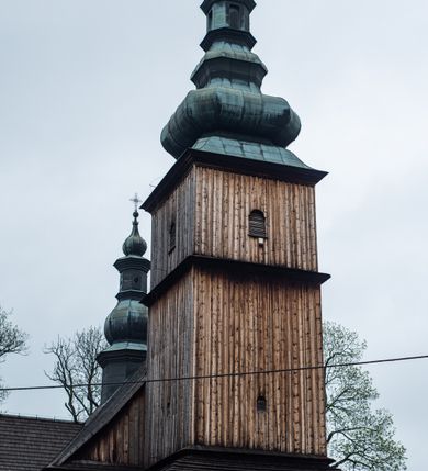 Zdjęcie nr 32: Kościół drewniany o konstrukcji zrębowej, elewacje jednolite, szalowane z listwowaniem, wzmocnione lisicami, wieża o konstrukcji słupowej. Więźba dachowa drewniana, krokwiowa. Budynek na kamiennym podmurowaniu przesłoniętym daszkiem gontowym. Kościół orientowany, jednonawowy z dwiema kaplicami o charakterze transeptu zamkniętymi wielobocznie, na planie krzyża łacińskiego, o węższym prezbiterium zamkniętym trójbocznie. Do wschodniej ściany prezbiterium przylega ogrójec w formie kapliczki, od północy zakrystia na planie prostokąta. Na skrzyżowaniu nawy z południowym ramieniem transeptu kruchta na planie prostokąta. Od zachodu do nawy przylega wieża na planie kwadratu, otoczona sobotami. Prezbiterium i nawa nakryte pozornym sklepieniem tej samej szerokości, obwiedzionym dużym, profilowanym gzymsem podtrzymywanym w nawie przez dwie pary filarów o kanelowanych trzonach, na cokołach. Sklepieniu nawy głównej towarzyszą płaskie i niższe stropy z fasetami w przestrzeniach bocznych. Ściany boczne nawy zwieńczone profilowanym gzymsem. Pomiędzy nawą a prezbiterium otwór tęczowy w formie odcinkowej arkady podtrzymywanej przez filary o kanelowanych trzonach z belką o falistym zarysie. Kaplice boczne otwarte do nawy odcinkowymi arkadami podtrzymywanymi przez filary o kanelowanych trzonach, na cokołach. Parapet chóru muzycznego w zachodniej części nawy, o wybrzuszonej środkowej części, podparty na dwóch filarach o kanelowanych trzonach, na cokołach. Posadzka z płyt ceramicznych. Elewacja frontowa w postaci wieży otoczonej sobotami. Na osi frontowej ściany sobót płytka kruchta zwieńczona gzymsem z przyczółkiem wypełnionym motywem „słoneczka”, przykryta dwuspadowym dachem. Drzwi dwuskrzydłowe, drewniane z okuciami. Korpus wieży podzielony poziomo daszkiem okapowym w dwóch trzecich wysokości, na froncie okno. W dolnej części małe otwory dzwonne z trzech stron, w górnej części duże otwory dzwonne z czterech stron. Wieża zwieńczona hełmem baniastym z latarnią. Ściany sobót szalowane z poziomym listwowaniem, przekryte mansardowym dachem gontowym, na północnej i południowej ścianie dwie pary okien w formie leżącego prostokąta zamkniętego łukiem odcinkowym. W elewacji południowej cztery otwory okienne w formie stojącego prostokąta zamkniętego łukiem odcinkowym, w partii prezbiterium z witrażami. Na elewacji wschodniej ogrójec w formie nadwieszonej kapliczki nakrytej dwuspadowym daszkiem z grupą Ukrzyżowania. W elewacji północnej dwa otwory okienne w formie stojącego prostokąta zamkniętego łukiem odcinkowym. W zakrystii małe kwadratowe okienko. W obu ramionach transeptu analogiczne jak w elewacjach otwory okienne na wschodniej i zachodniej ścianie. Kościół nakryty dachami gontowymi, wyższym nad nawą, niższym nad prezbiterium, dwuspadowym nad nawą oraz wielospadowym nad prezbiterium i ramionami transeptu; nad zakrystią i kruchtą boczną dachy dwuspadowe. W połowie długości dachu, nad nawą, od strony prezbiterium wznosi się ponad kalenicą wieżyczka na sygnaturkę, obita blachą, na ośmiobocznym trzonie, z latarnią zwieńczoną niewielkim baniastym hełmem z latarnią.

